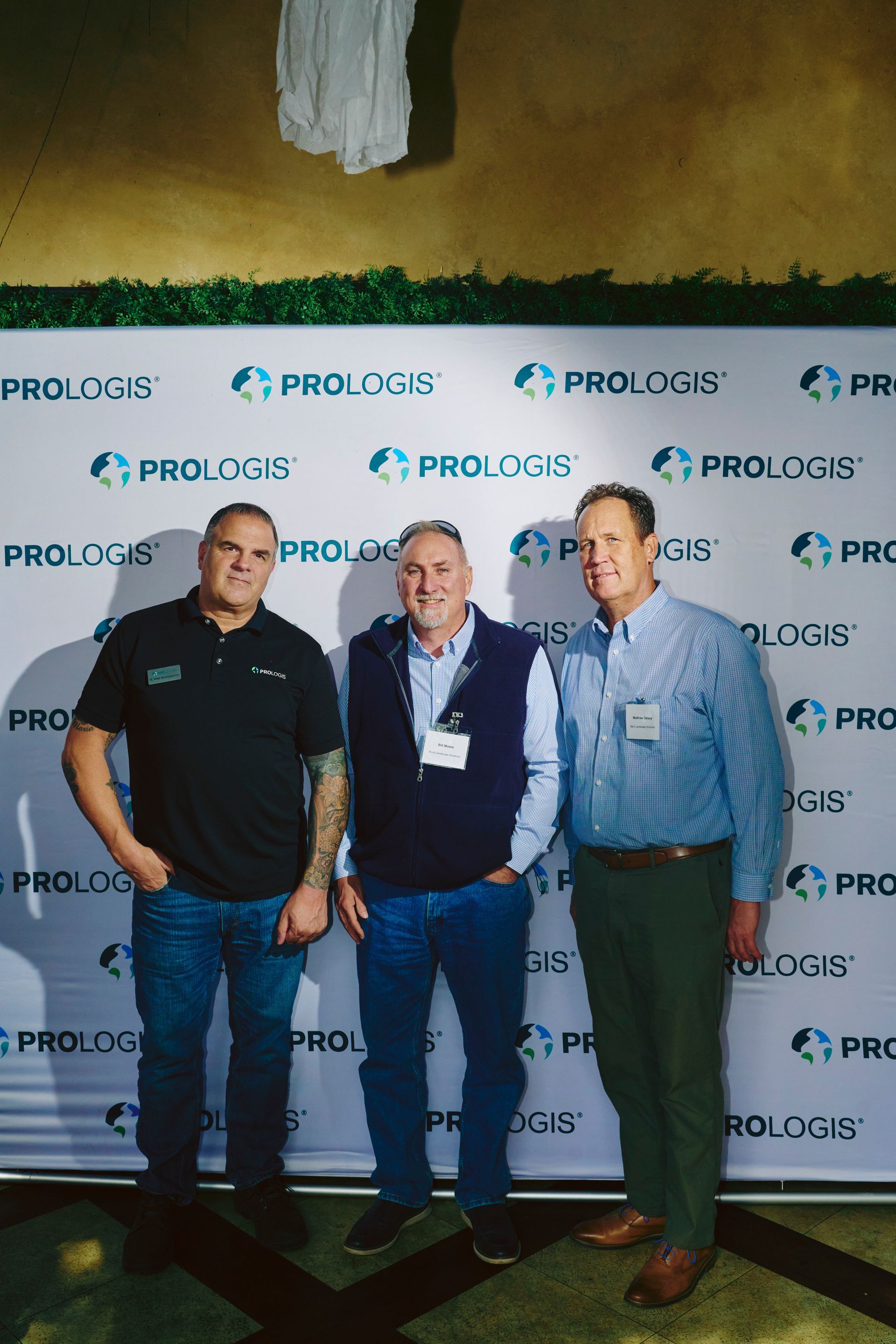 Three men pose in front of a backdrop with the company name, Prologis. Two are wearing vests, and one is wearing a dark t-shirt.