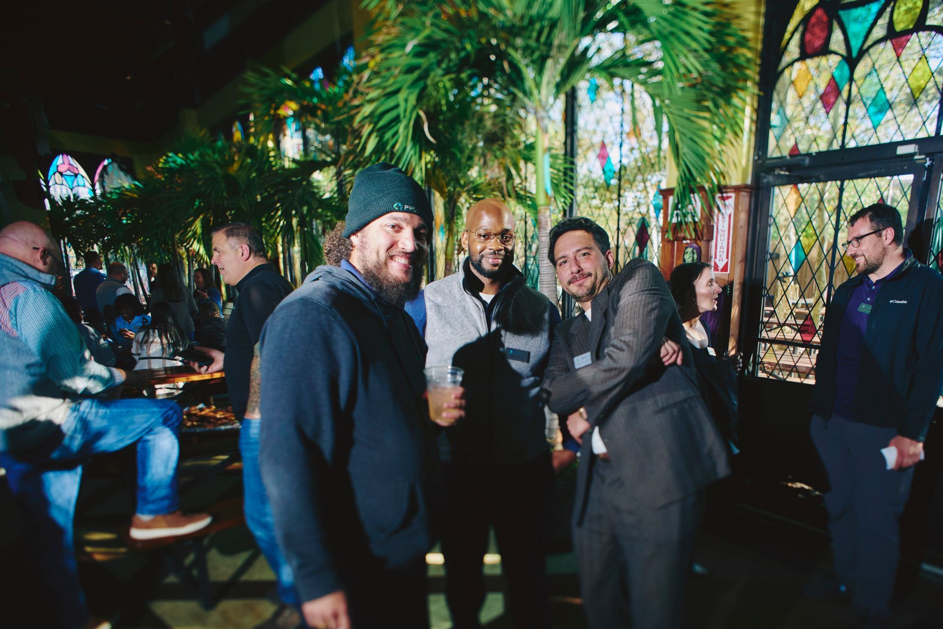 Three men posing together indoors near stained glass windows, smiling. Other people visible in the background.