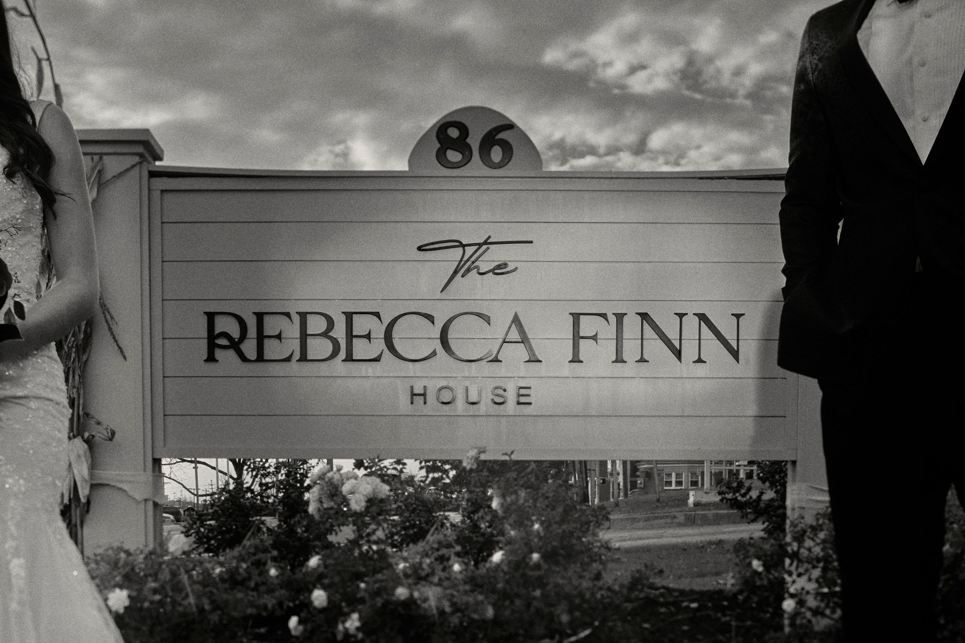 A black and white photo of a bride and groom standing in front of a sign for the rebecca finn house.