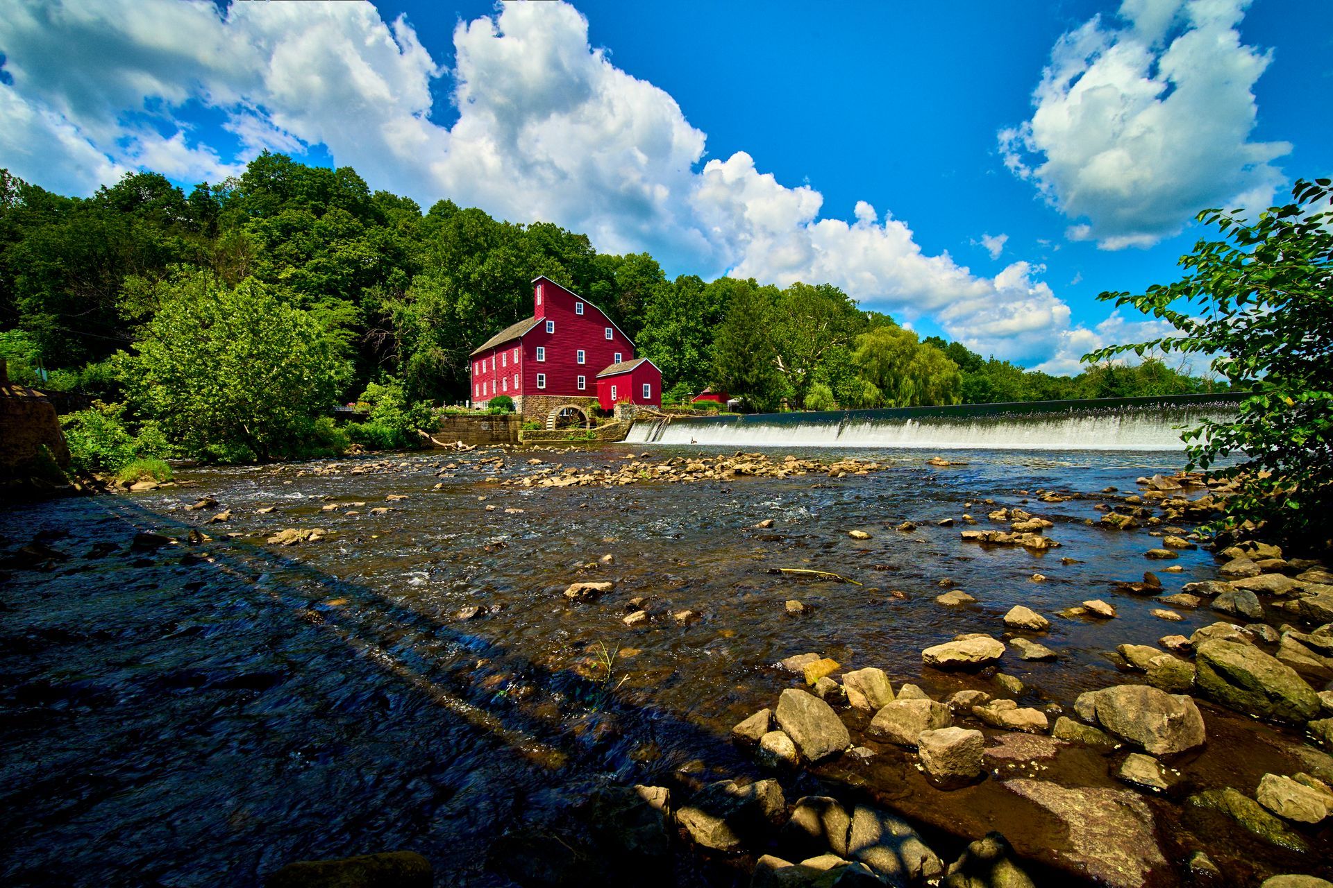 Red mill building next to a rocky river under a blue sky with fluffy white clouds.