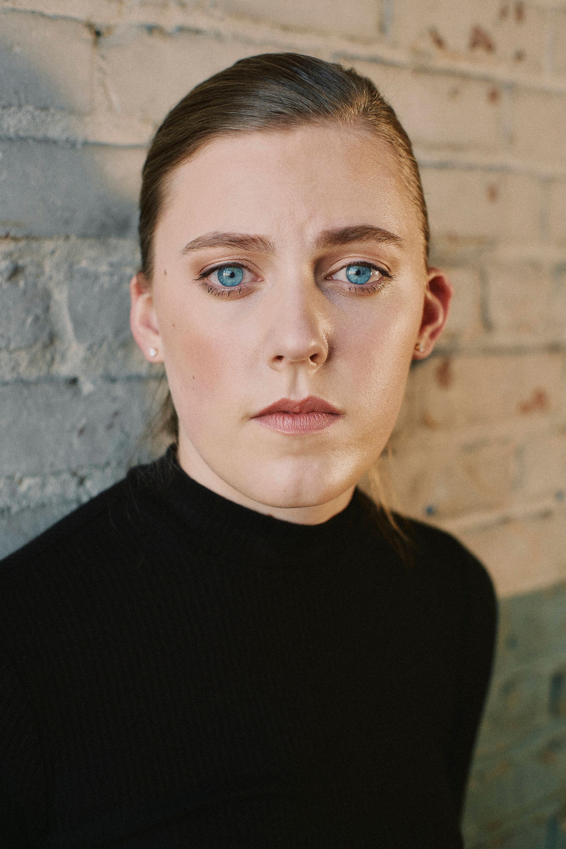 A woman with blue eyes and a black turtleneck is standing in front of a brick wall.