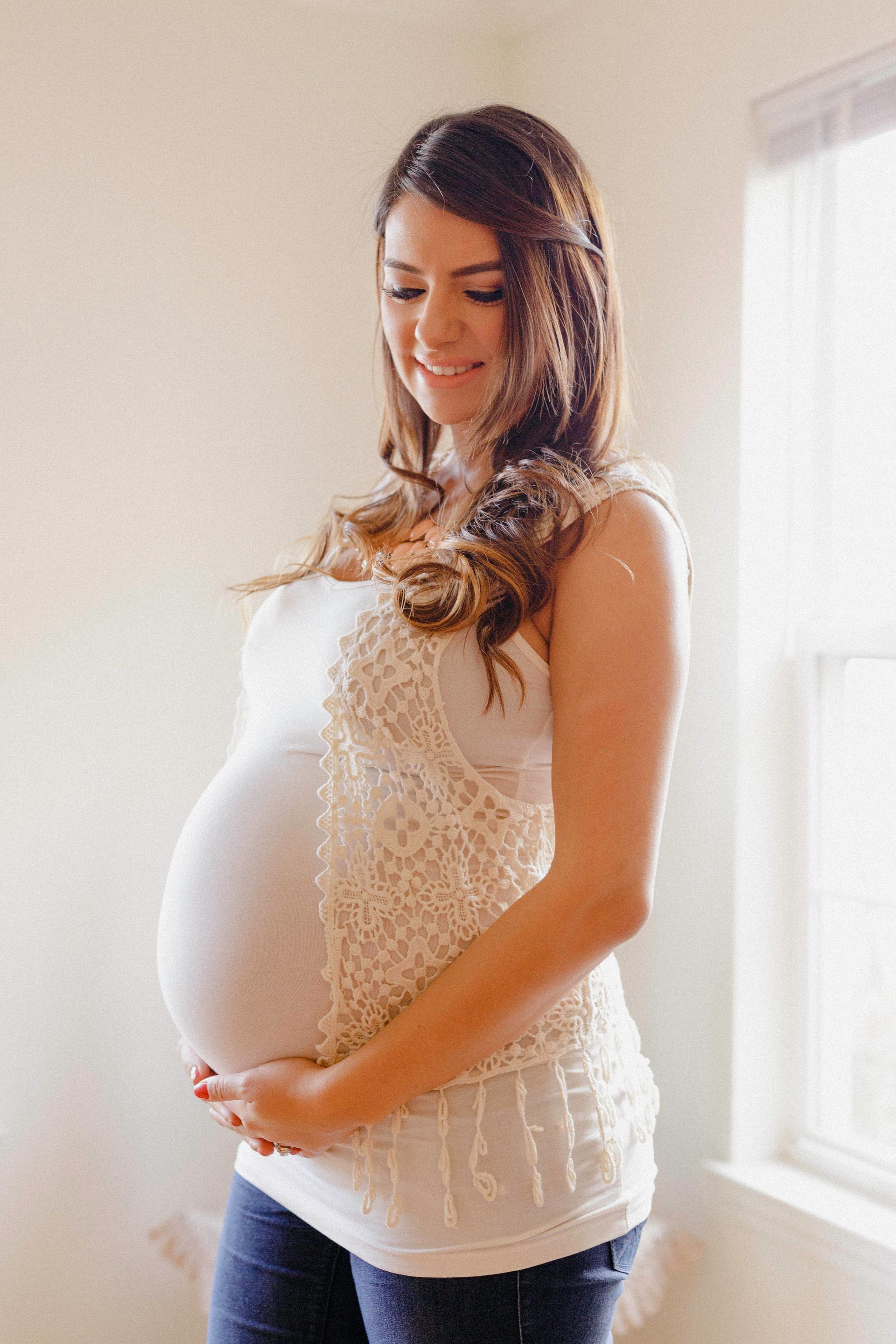 Pregnant person holding belly, smiling, wearing white top and jeans, near a window.