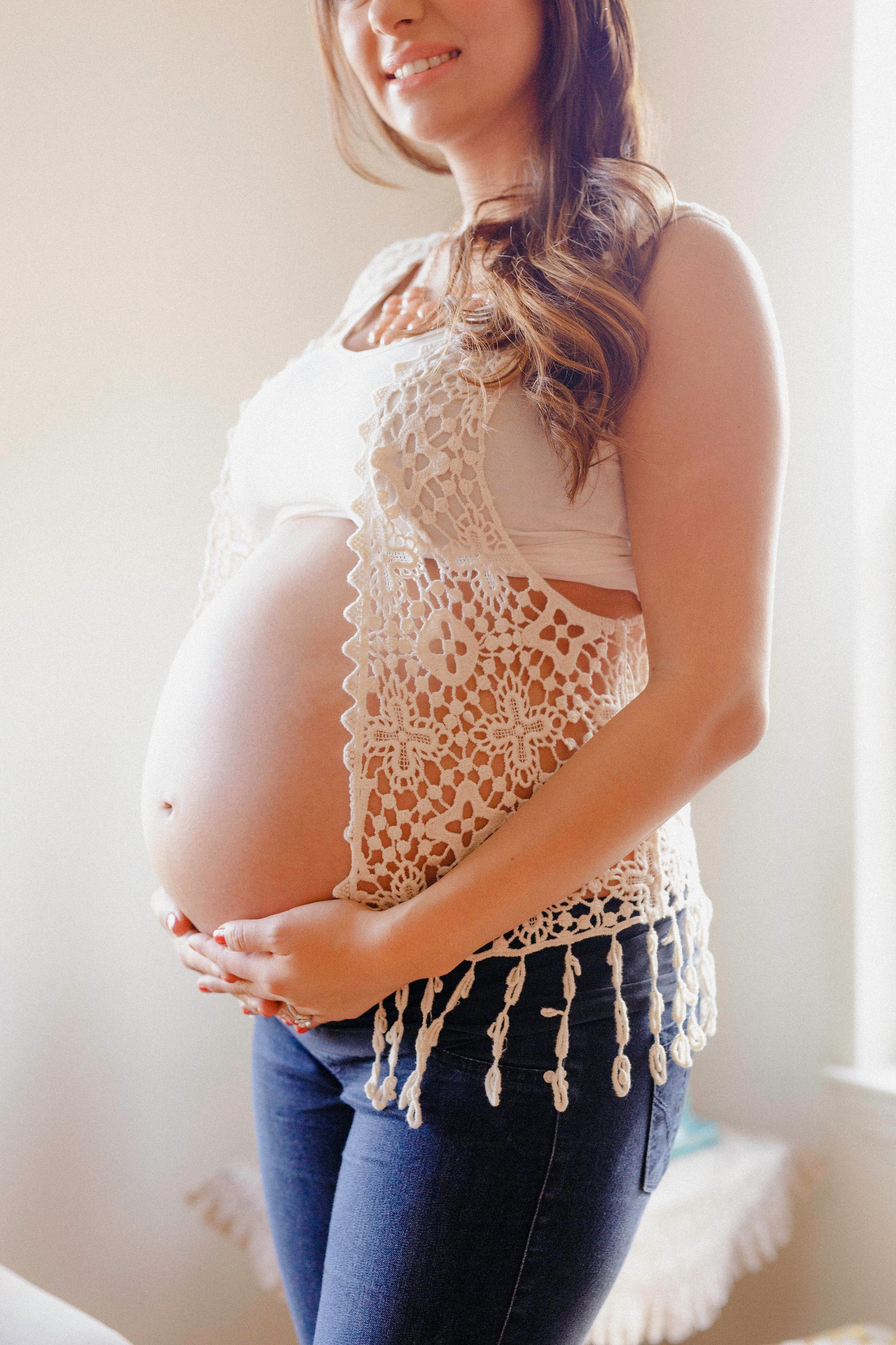 A pregnant woman is standing in front of a window holding her belly.