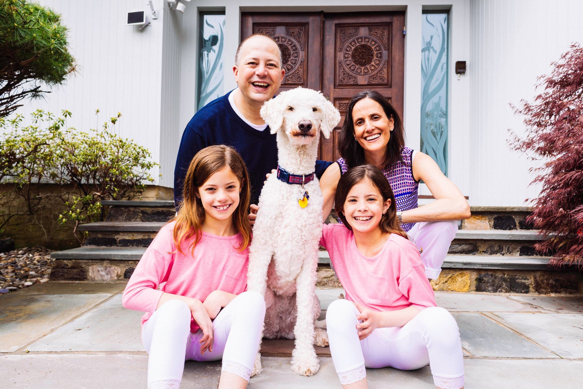 Family of four with a dog, posing in front of their home. Two young girls in pink, parents smiling.