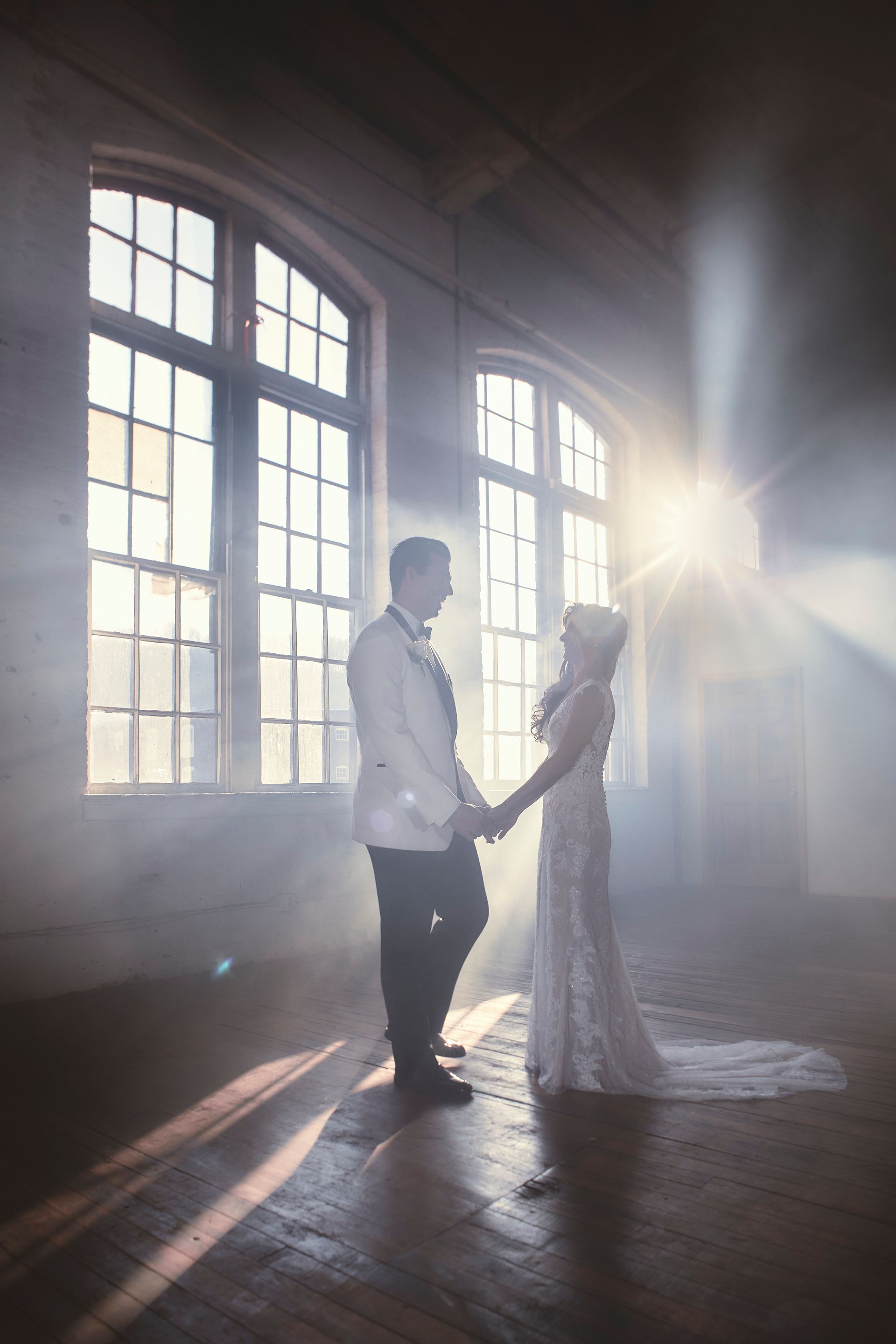 A bride and groom are standing in front of a window holding hands.
