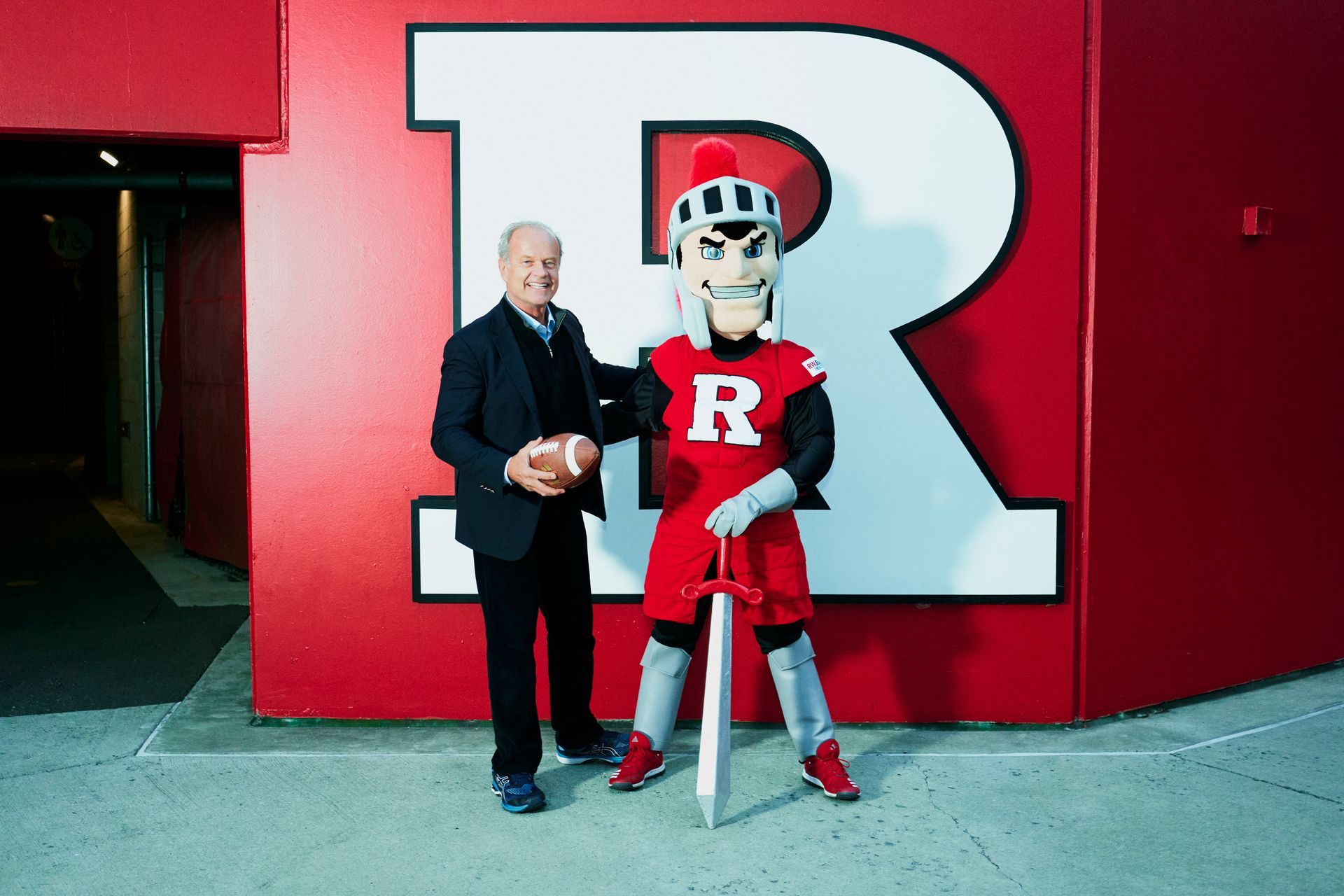 A man and a mascot are standing in front of a large letter r