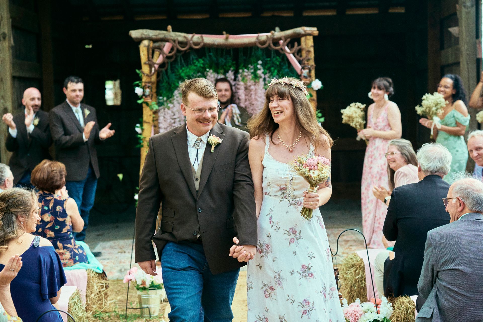 Newlyweds walk hand-in-hand down the aisle after a barn wedding. Guests clap, floral arch behind.