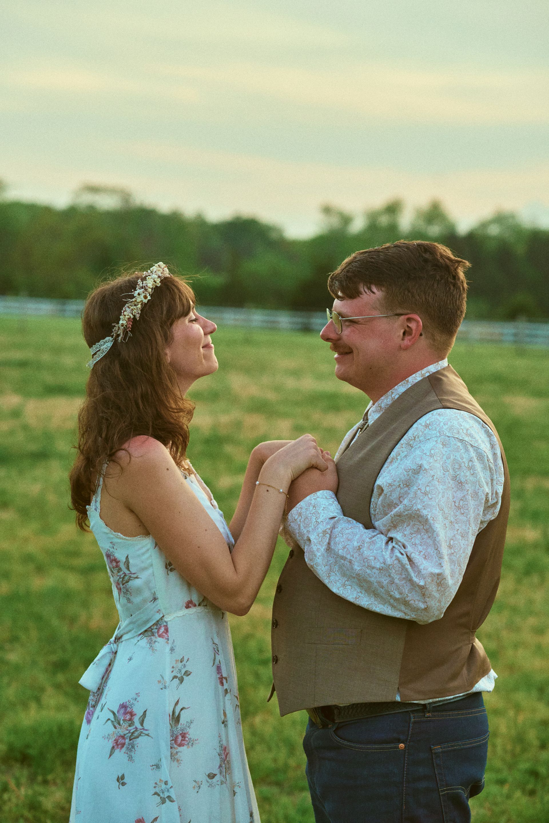 Couple holding hands, smiling at each other in a grassy field. Woman wears a floral dress and headband. Man wears a vest.