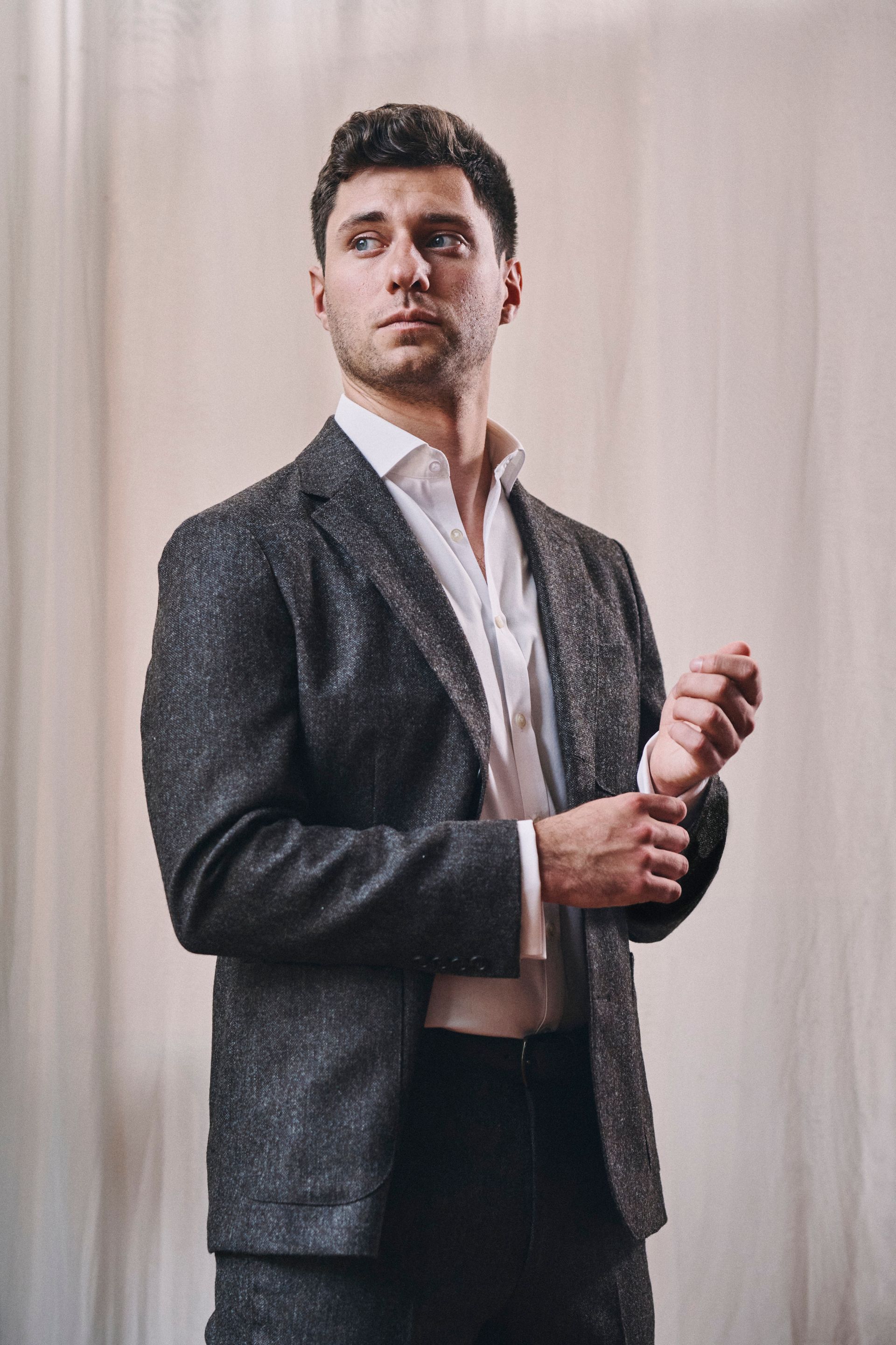 A man in a suit and white shirt is standing in front of a white curtain.