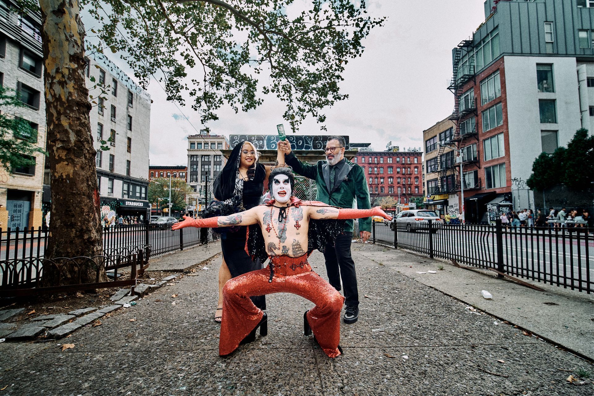 Person in red pants poses with arms outstretched on a gravel path in an urban street scene.
