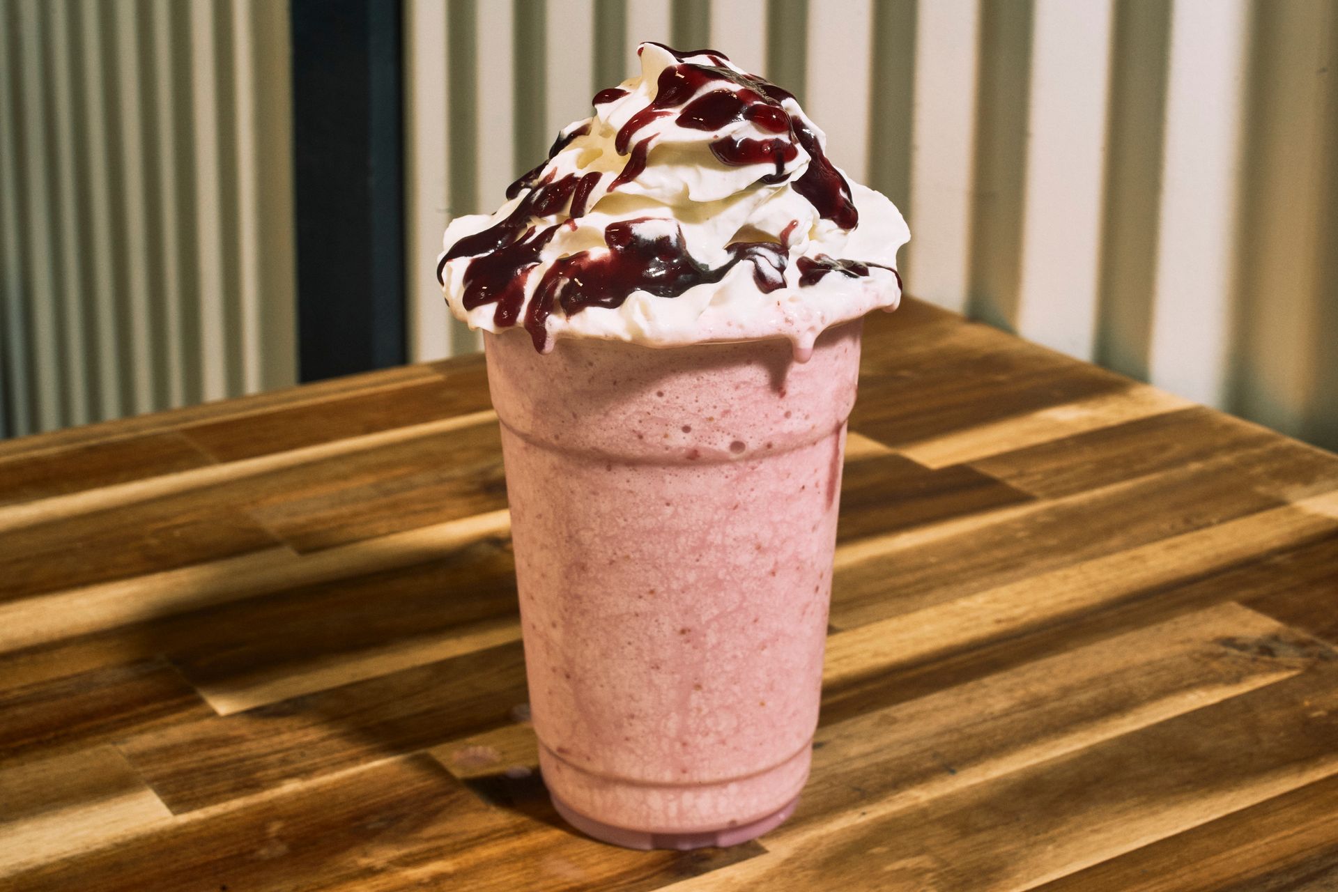 Purple milkshake in a clear cup, topped with whipped cream and berry drizzle on a wooden table.