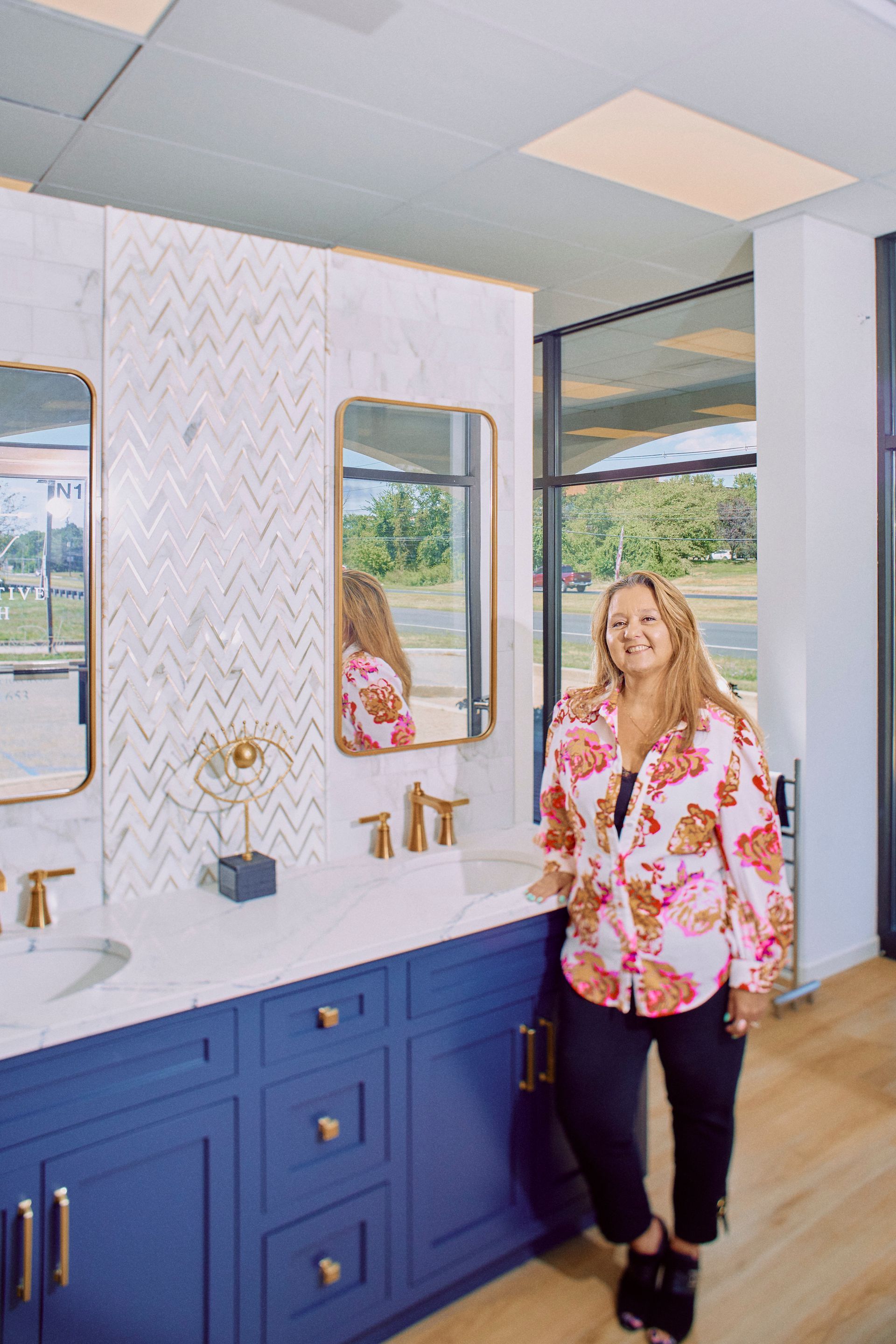 Woman in floral shirt stands by blue bathroom vanity in showroom.