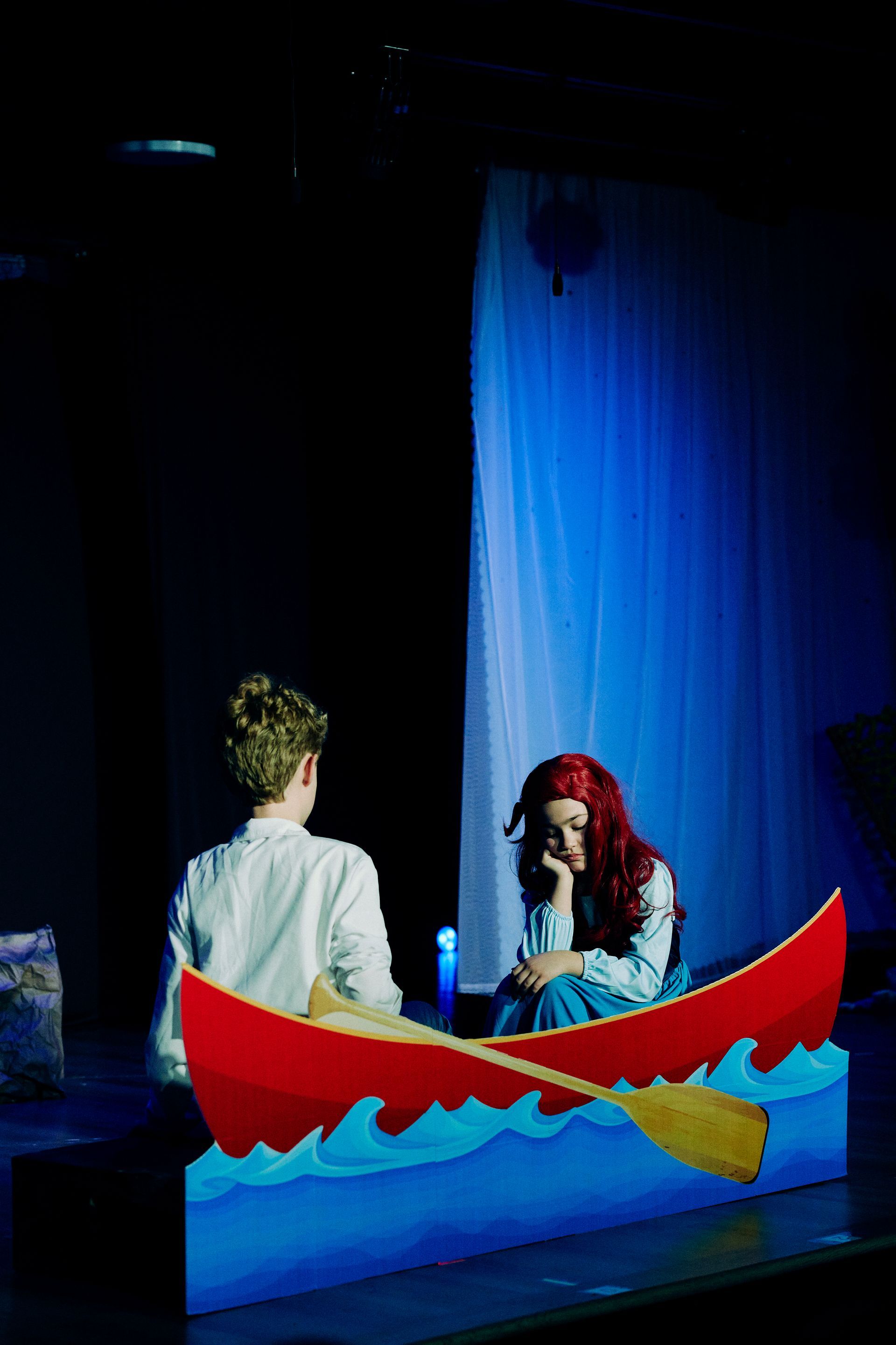 Two people in a small boat on a stage, with blue backdrop and bright red-and-yellow props