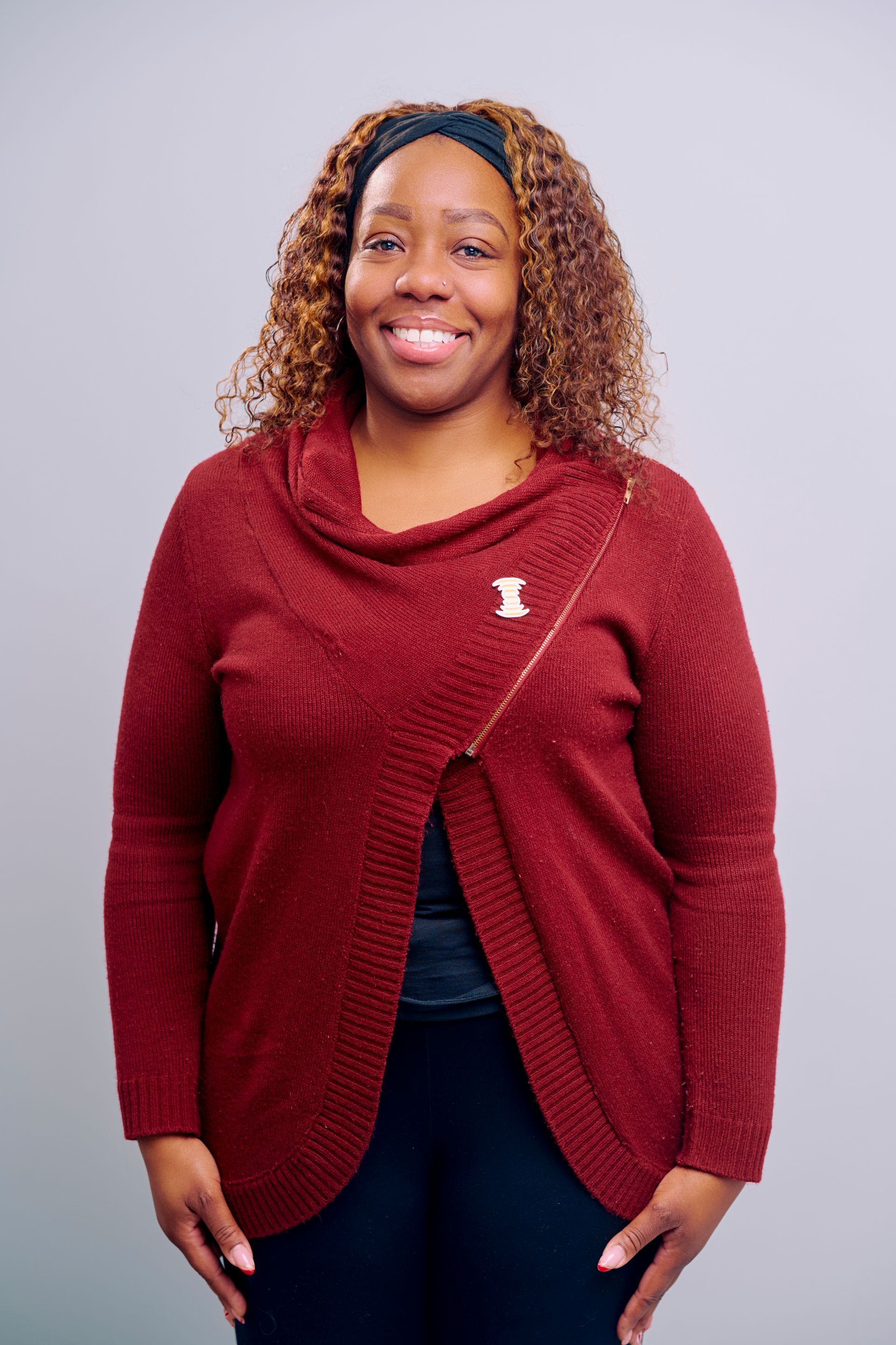 Woman smiling, wearing a burgundy sweater and black leggings, standing in front of a gray backdrop.