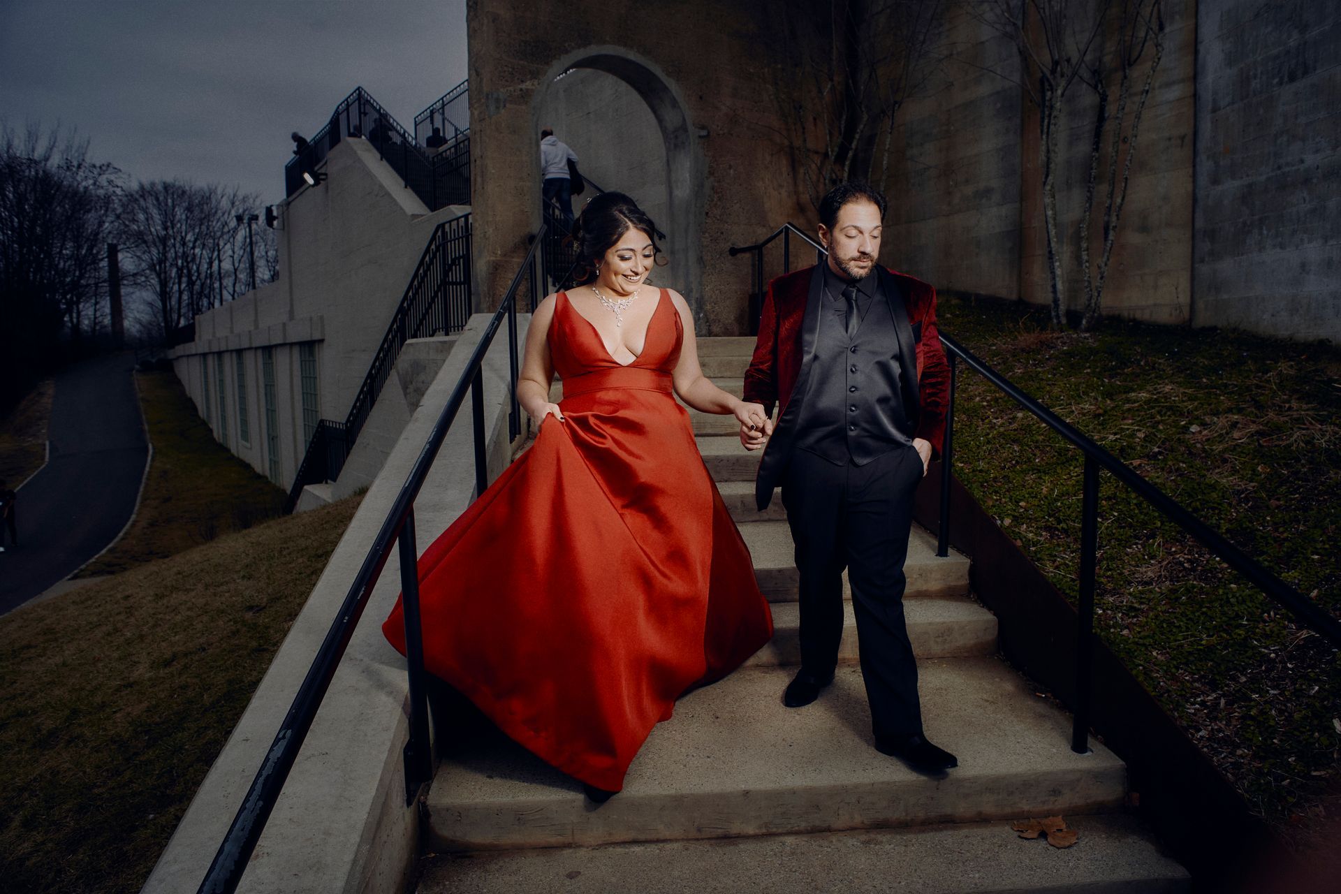 A man and a woman are walking down stairs holding hands . the woman is wearing a red dress.