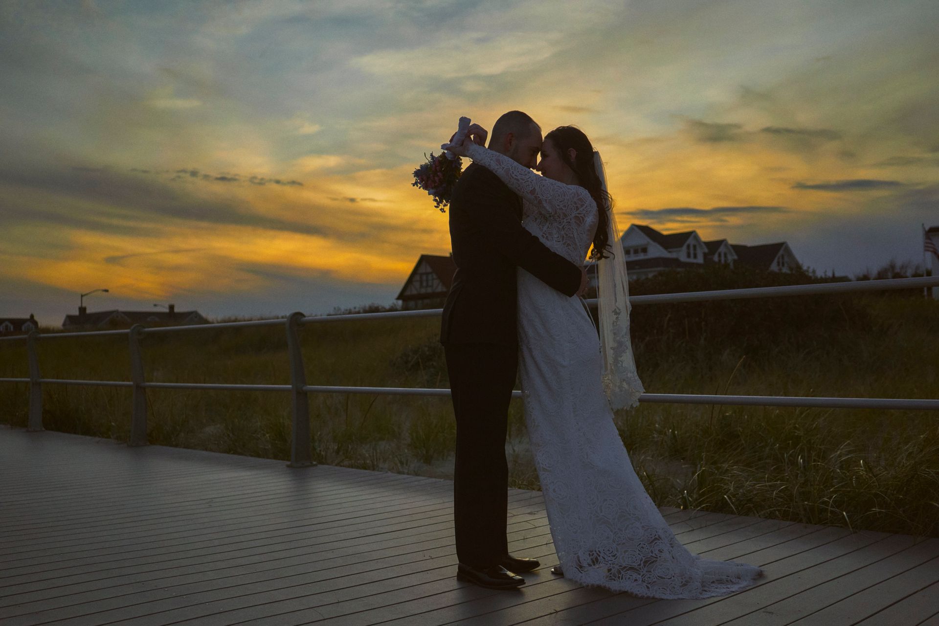 A bride and groom are kissing on a pier at sunset.