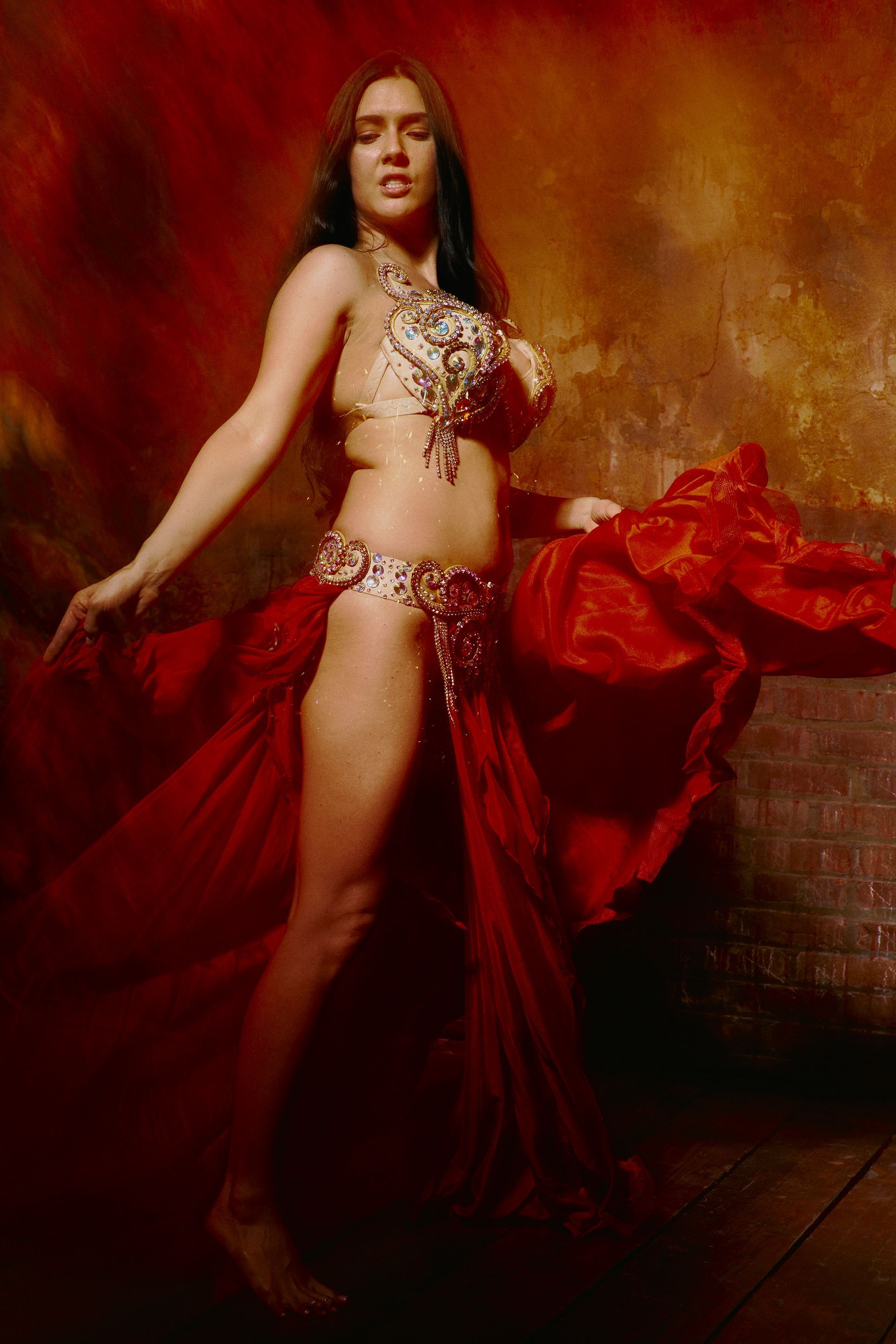 A belly dancer in a red dress is dancing in front of a wall.