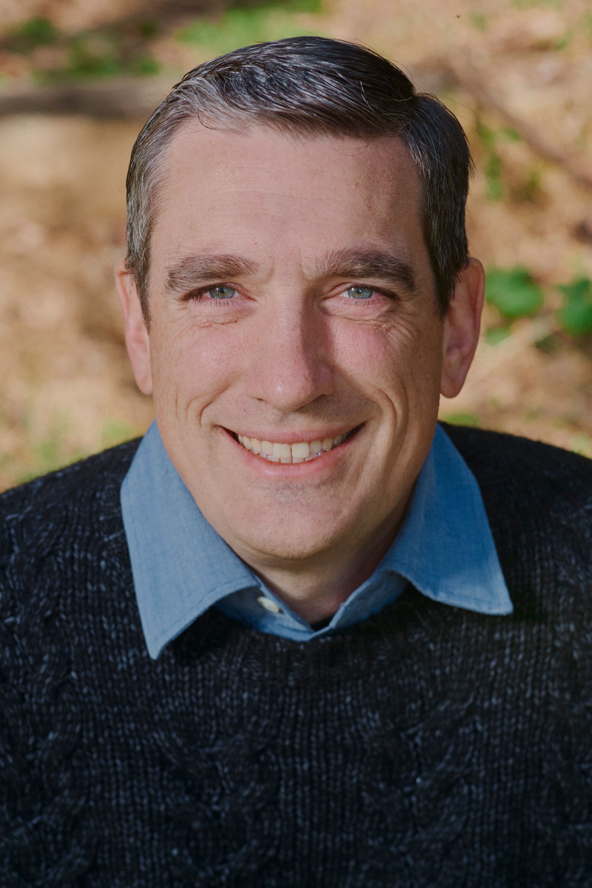 A man wearing a blue shirt and a black sweater smiles for the camera