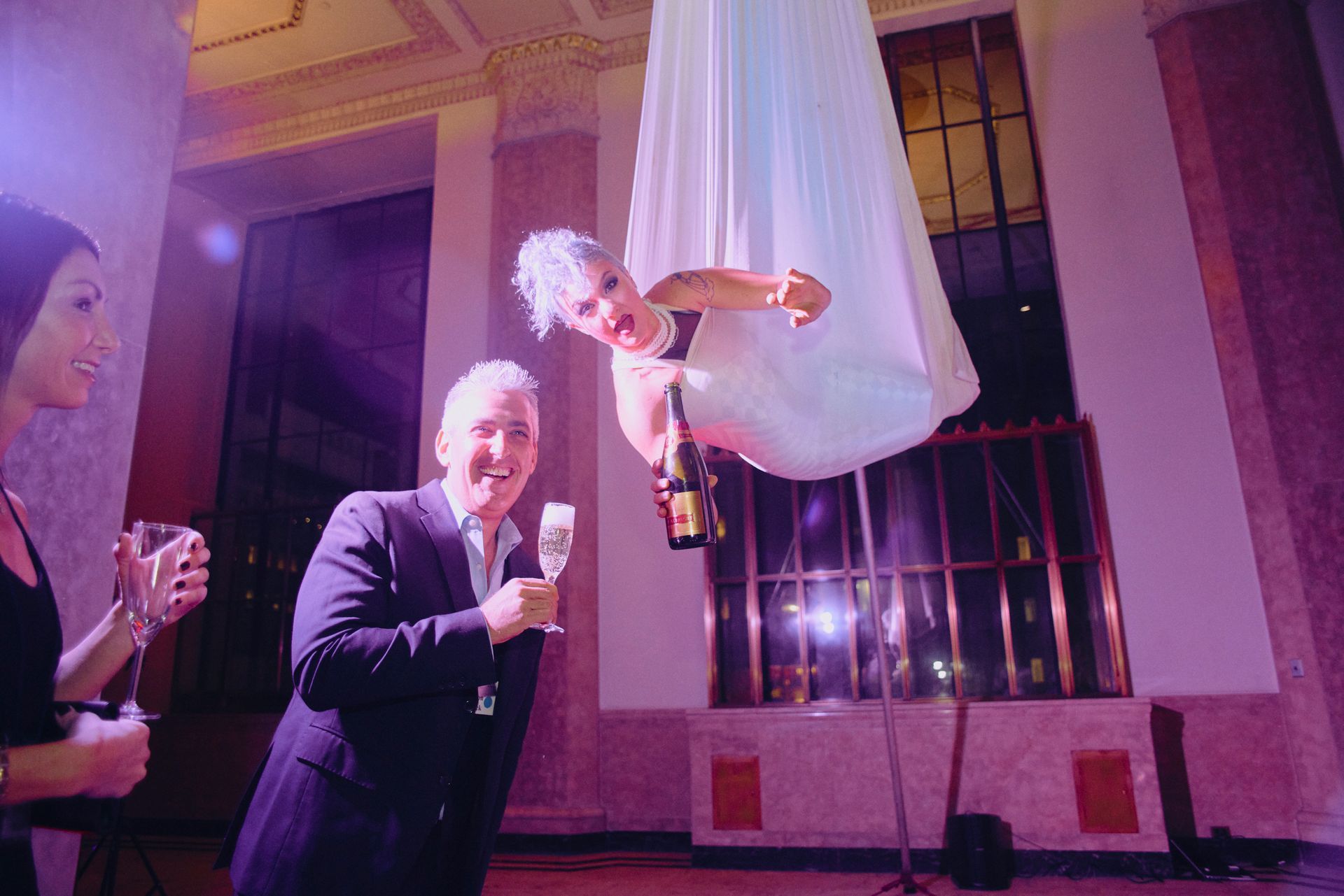 A woman in a white dress is hanging upside down in the air while holding a bottle of champagne.