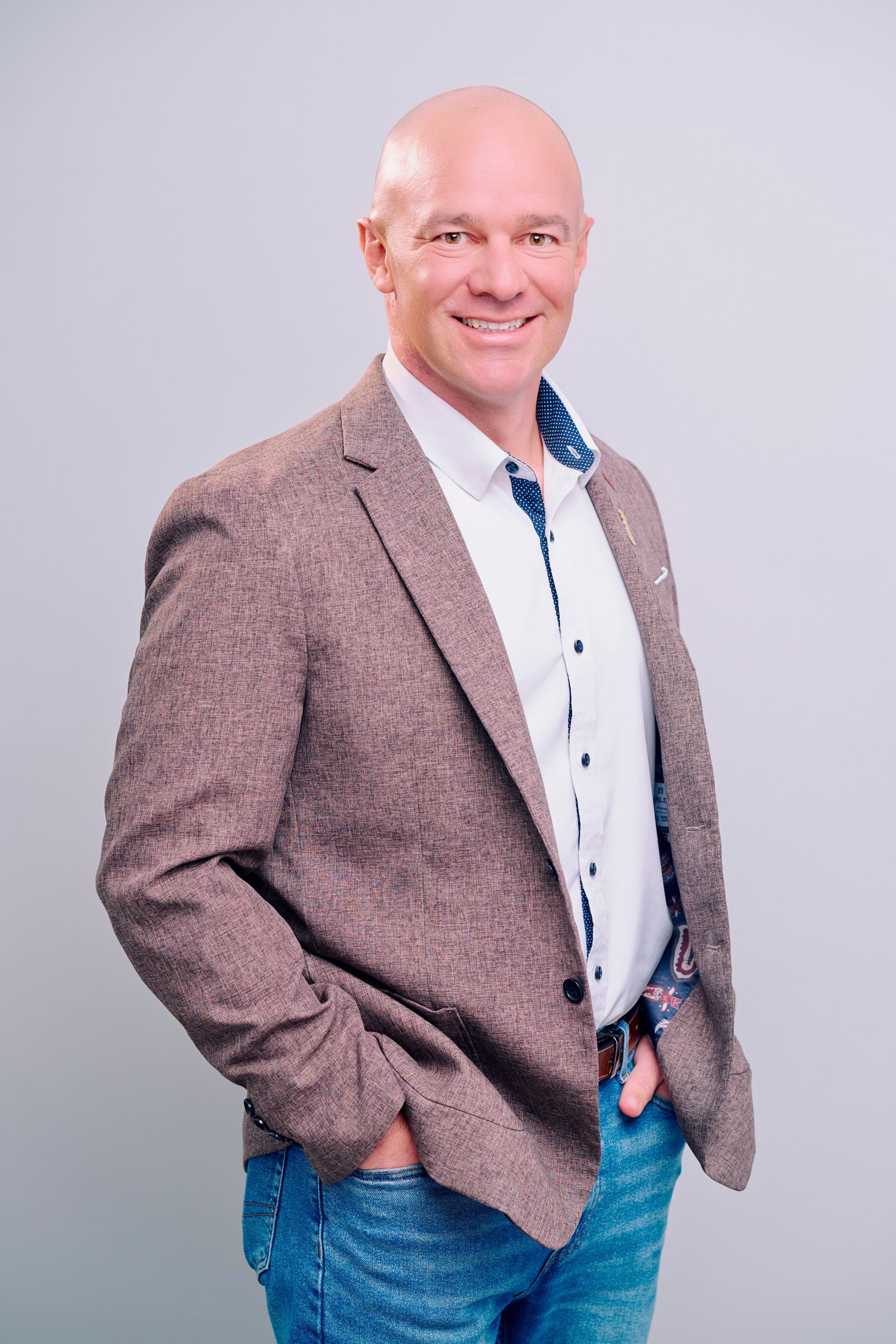Bald man in brown blazer and jeans smiles, hands in pockets.
