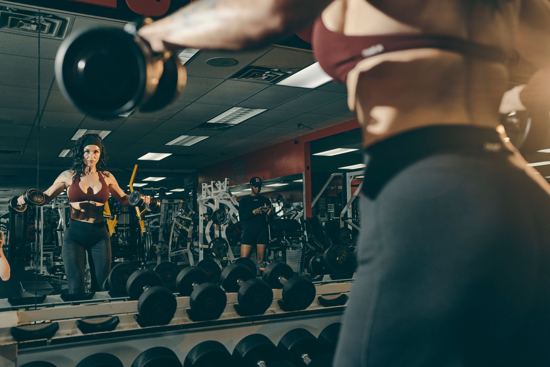Gym scene with a person lifting a dumbbell in front of a mirror and rows of weights