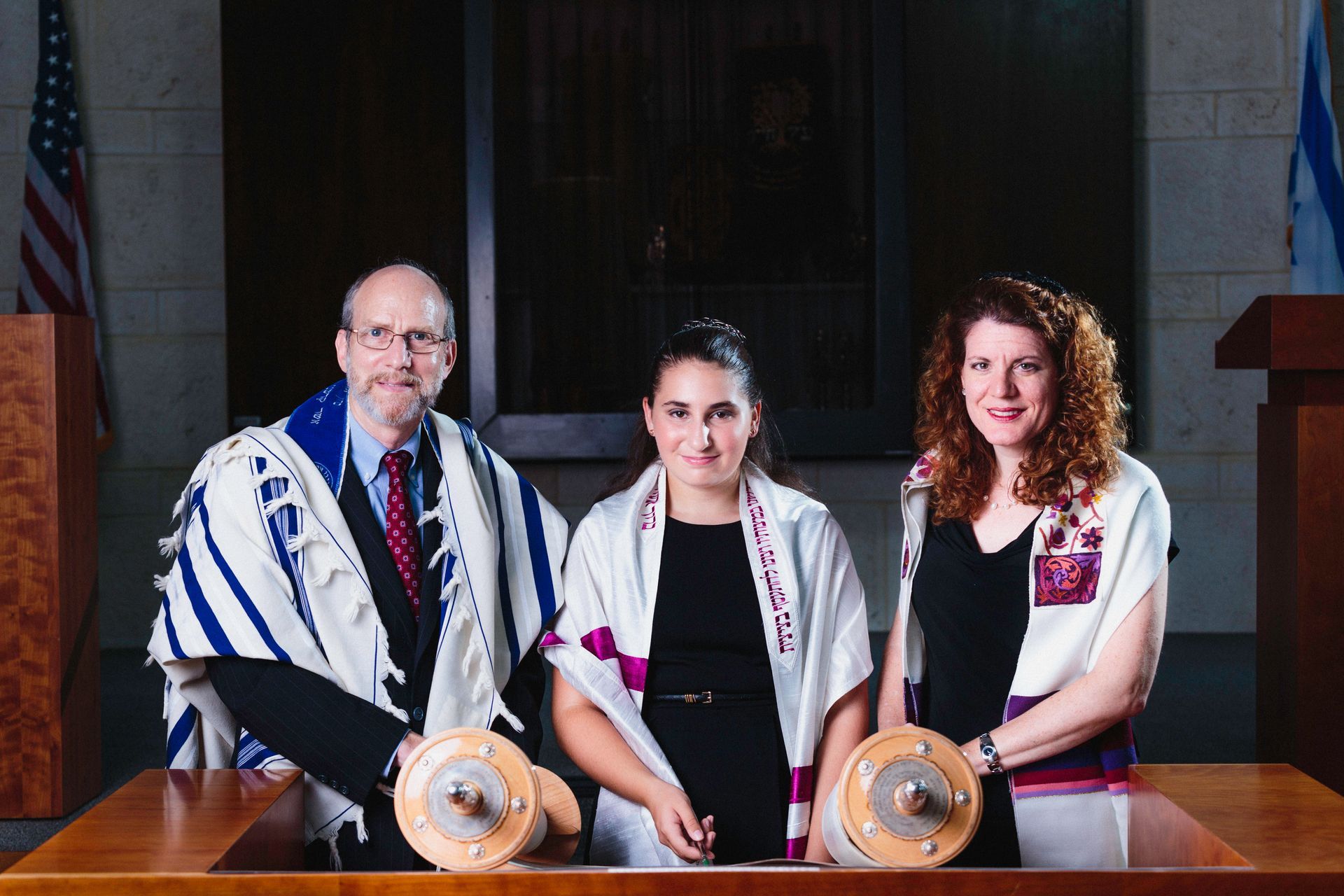 Three people with Torah scrolls at a synagogue. The middle person is smiling, draped in a prayer shawl. Flags in background.