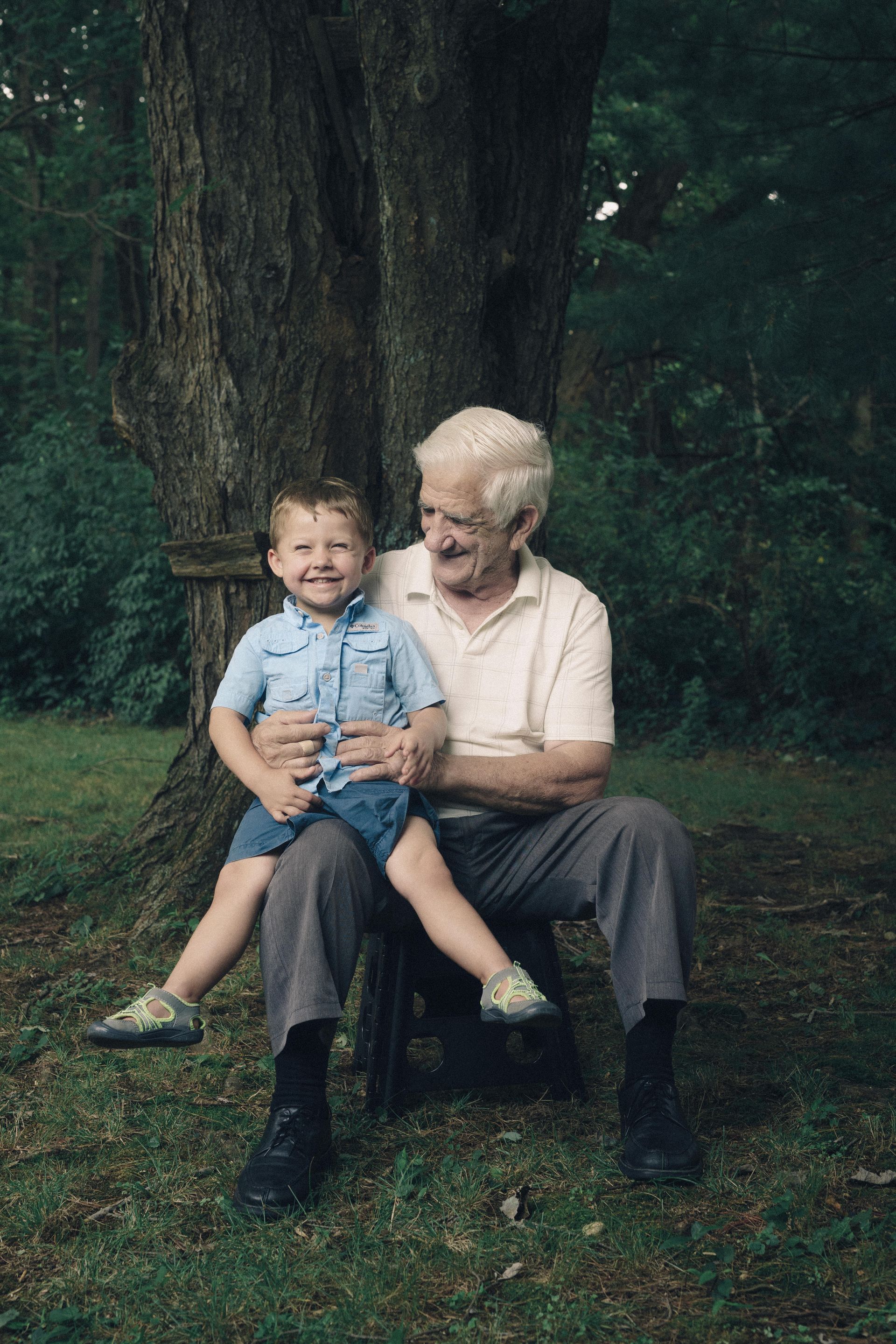 Grandfather sits outdoors, holding a smiling child on his lap. They are by a tree on grass.