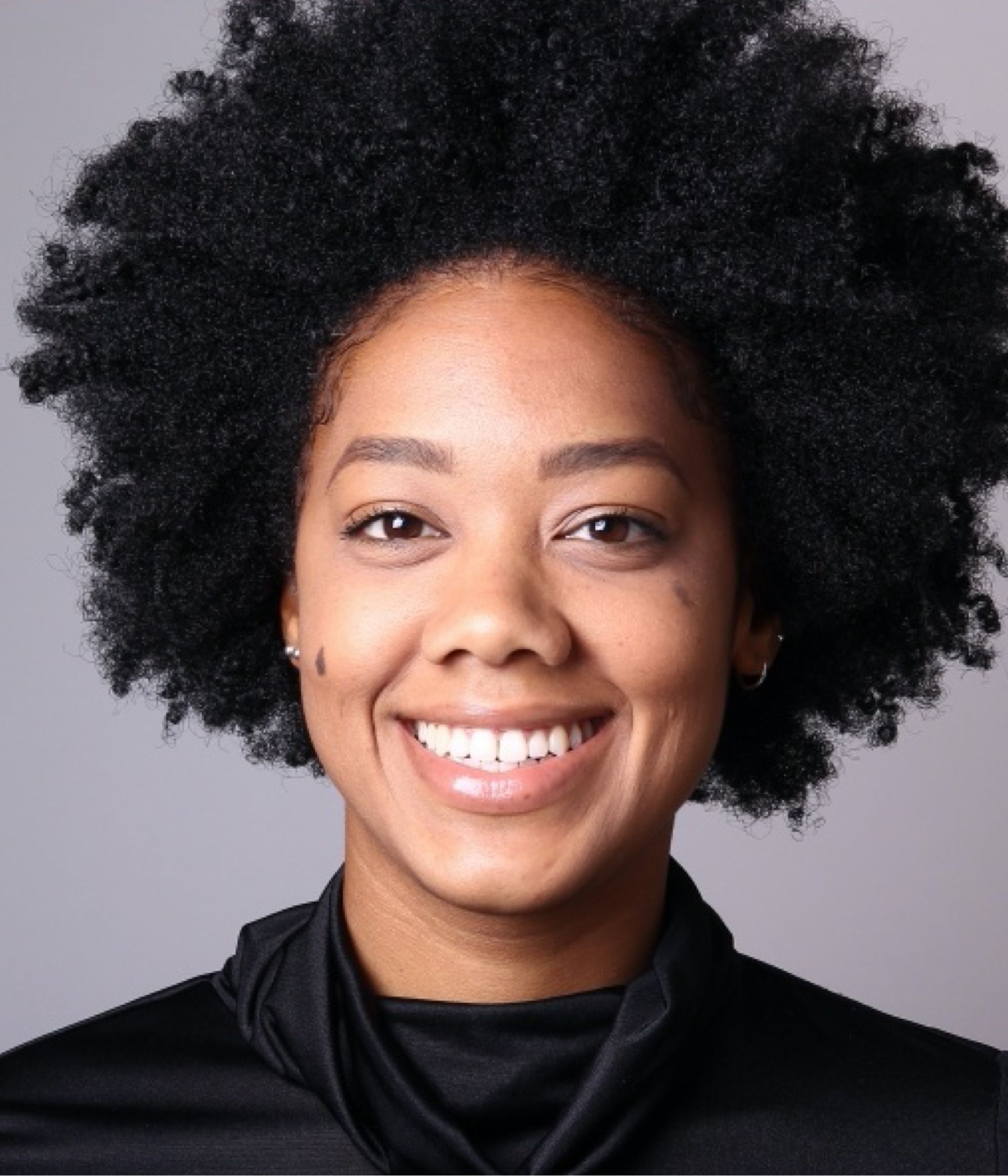 Person with curly hair smiles, wearing a black top against a gray backdrop.