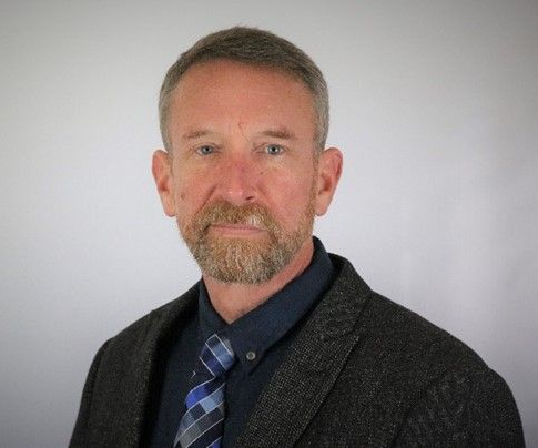 Peter Hitschler smiles, wearing a dark blazer and blue tie, in an office setting.