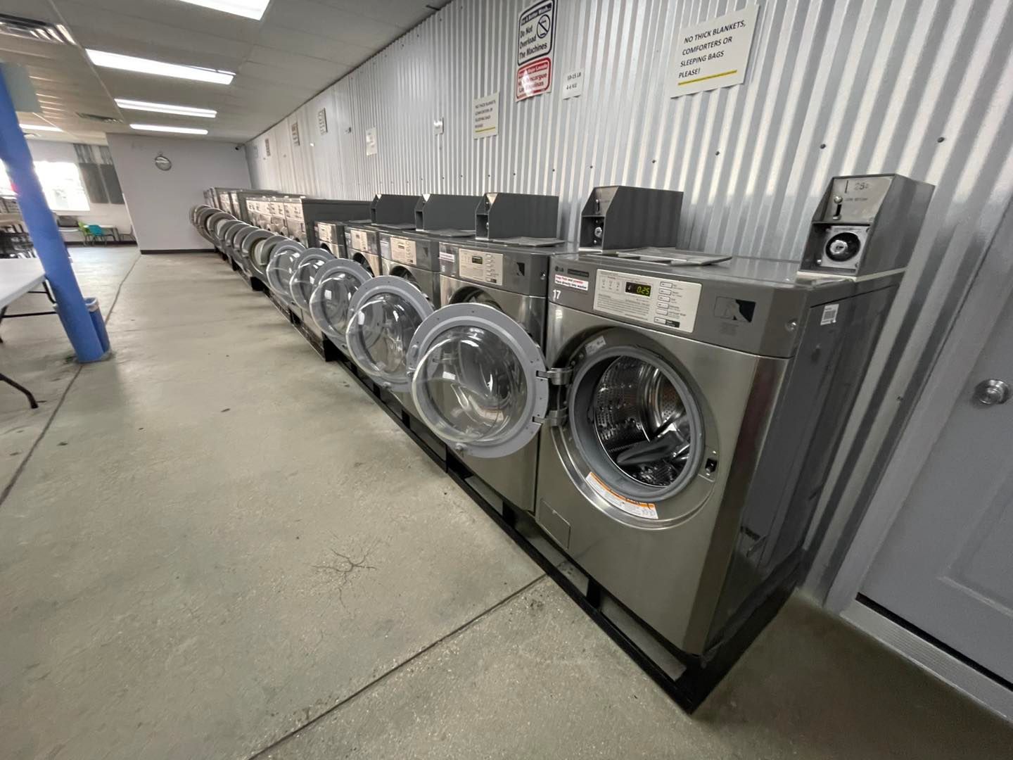 Row of front-loading washing machines in a laundromat with a concrete floor and metal wall