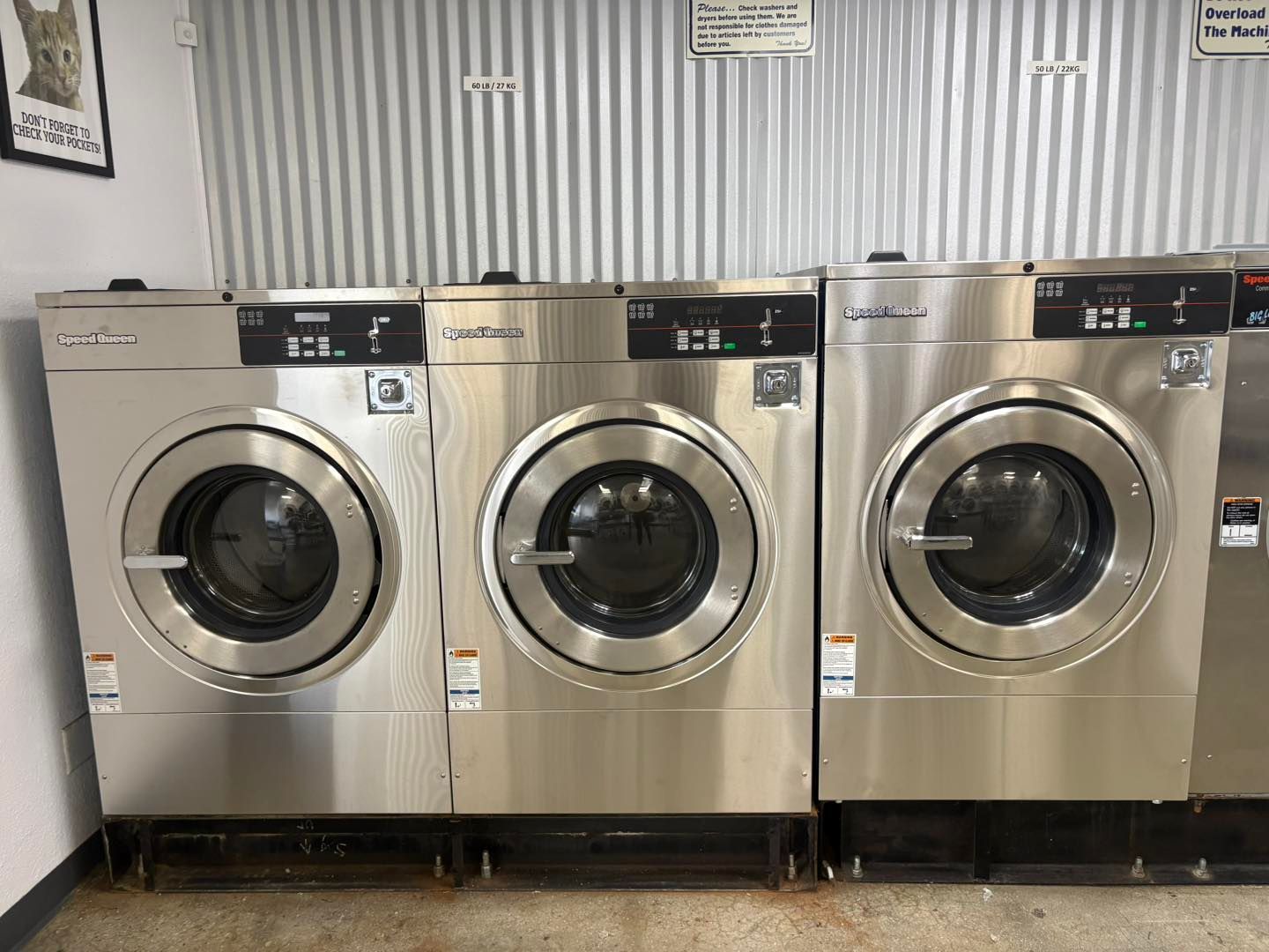 Three front-loading washing machines in a laundromat against a corrugated metal wall.