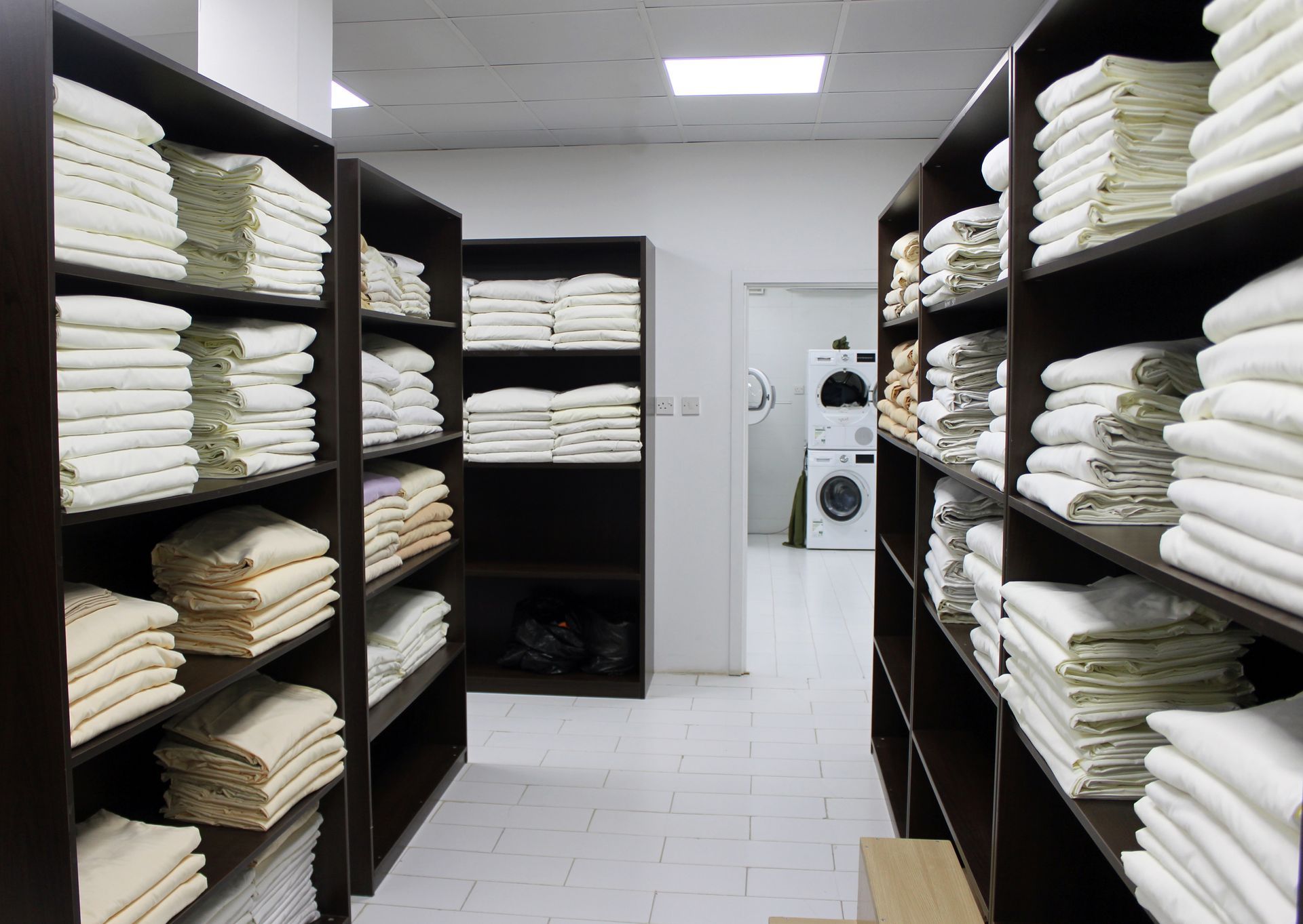 Shelves of folded white linens in a laundry storage room with washers in the back.
