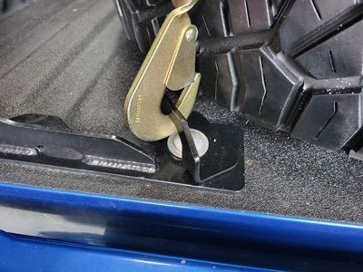 A metal hook securing a tire in a blue truck bed, attached to a black tie-down point.