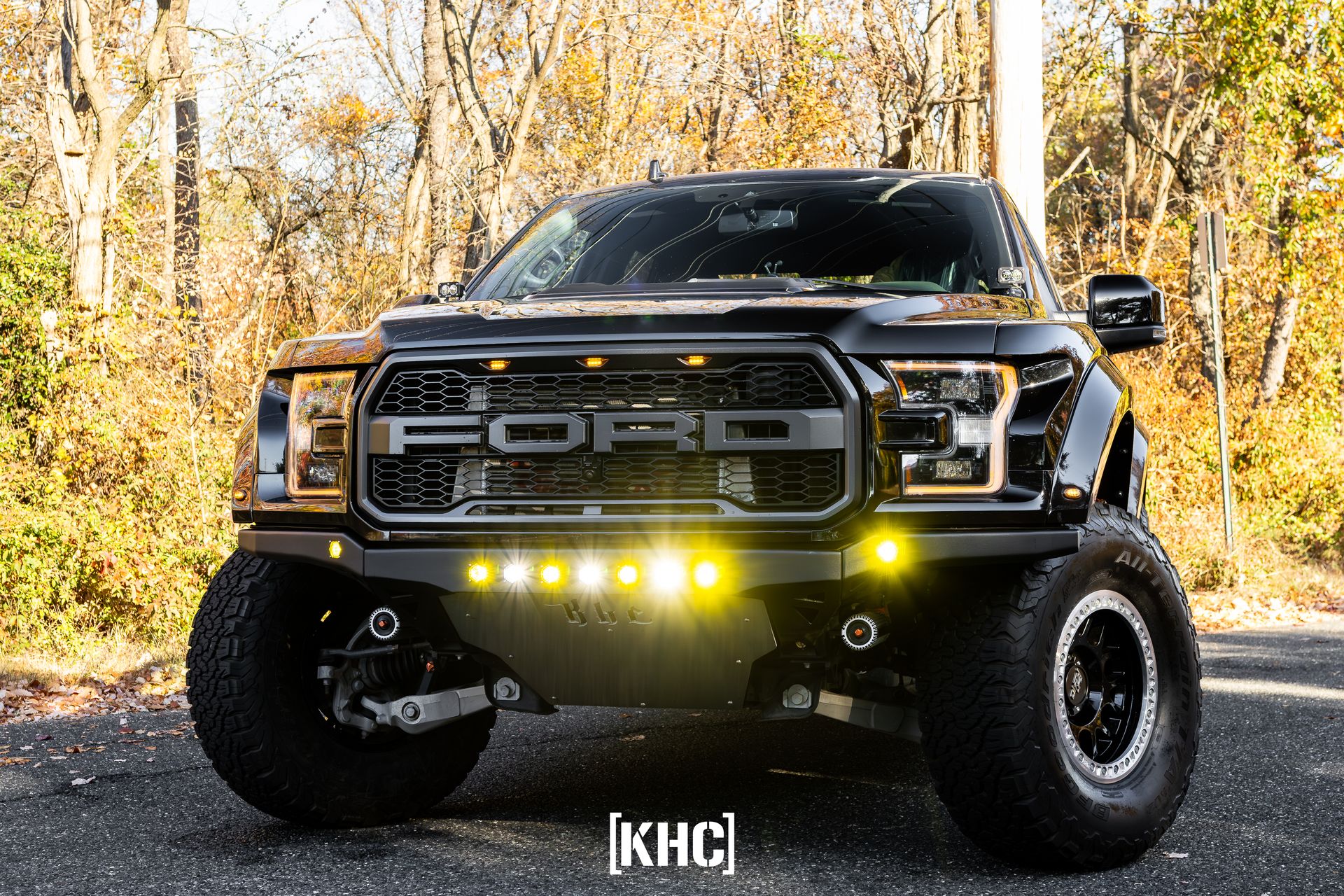 Black Ford Raptor truck with yellow lights, parked on a road with fall foliage in the background.