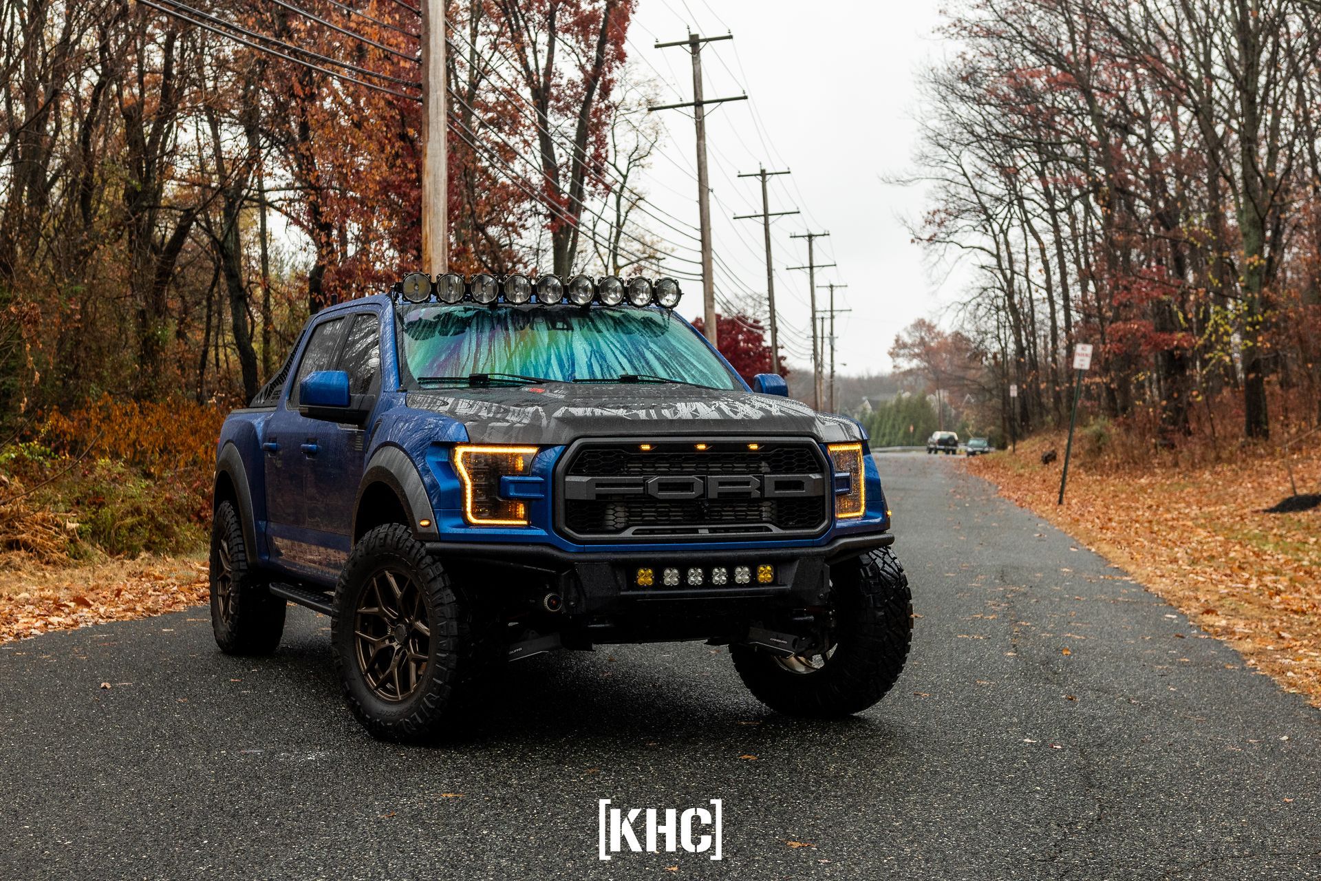 Blue Ford Raptor truck on a wet road, surrounded by autumn trees. Lights mounted on the roof and front bumper.
