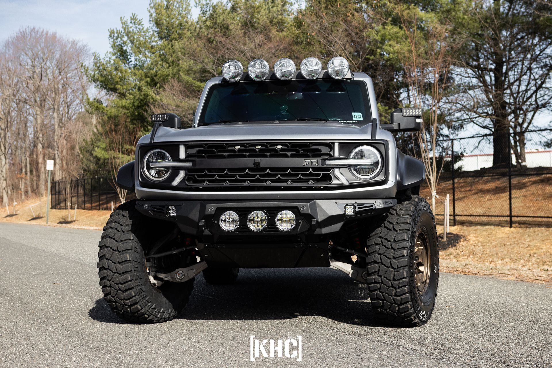 Gray, customized Ford Bronco with large tires, off-road lights, and a black grille, parked on pavement.