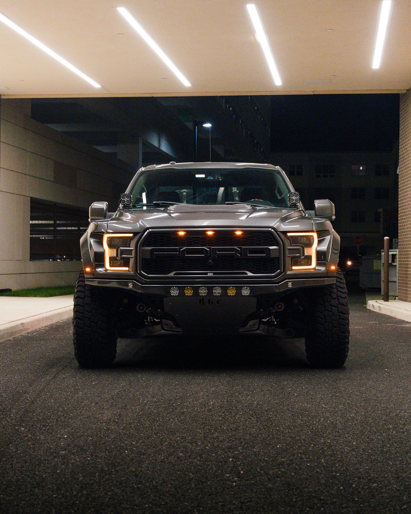 Gray Ford Raptor truck, front view, under illuminated overhang at night.