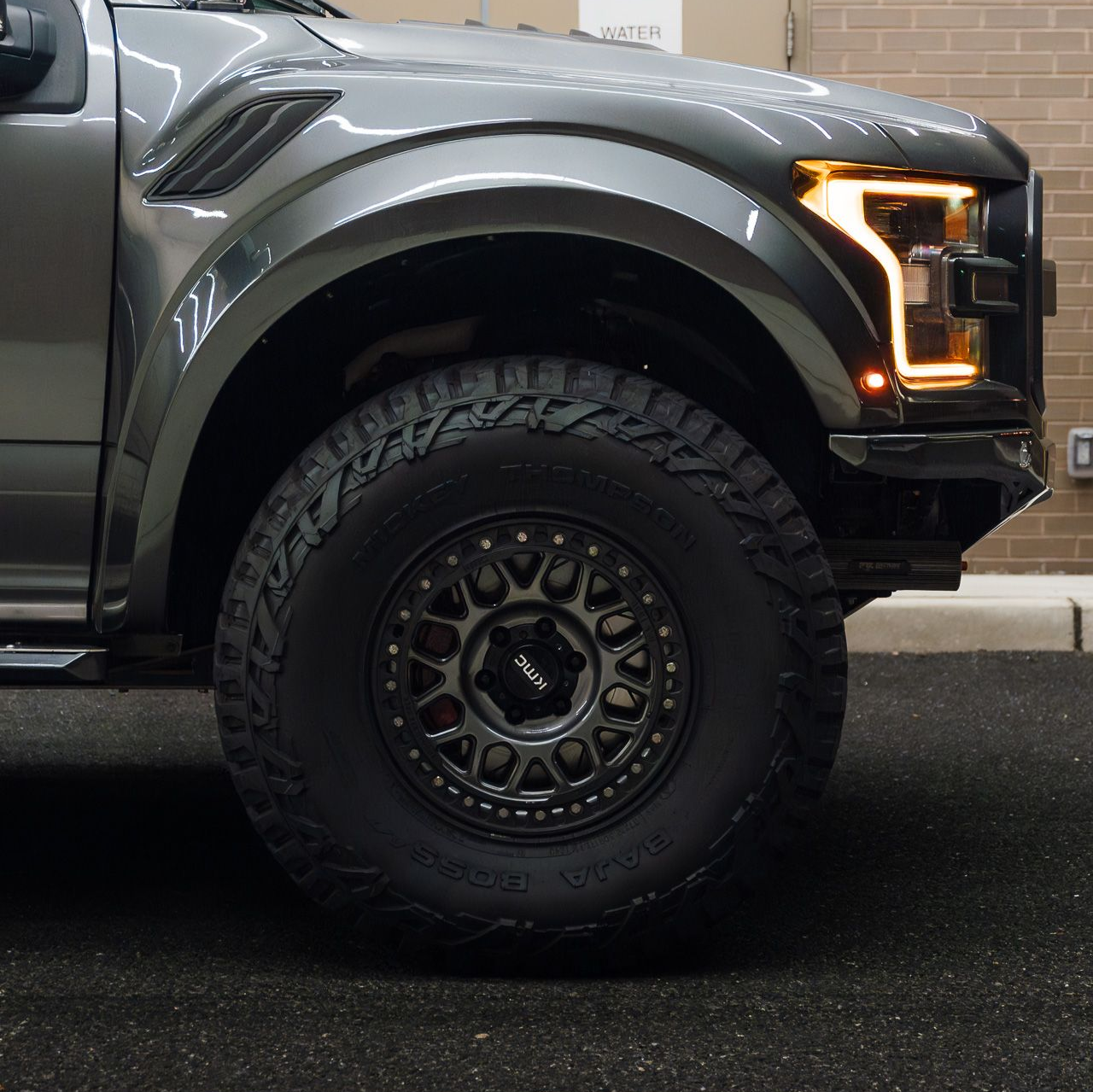 Gray Ford Raptor truck, front wheel and headlight visible, on asphalt. Black tire, off-road wheel.