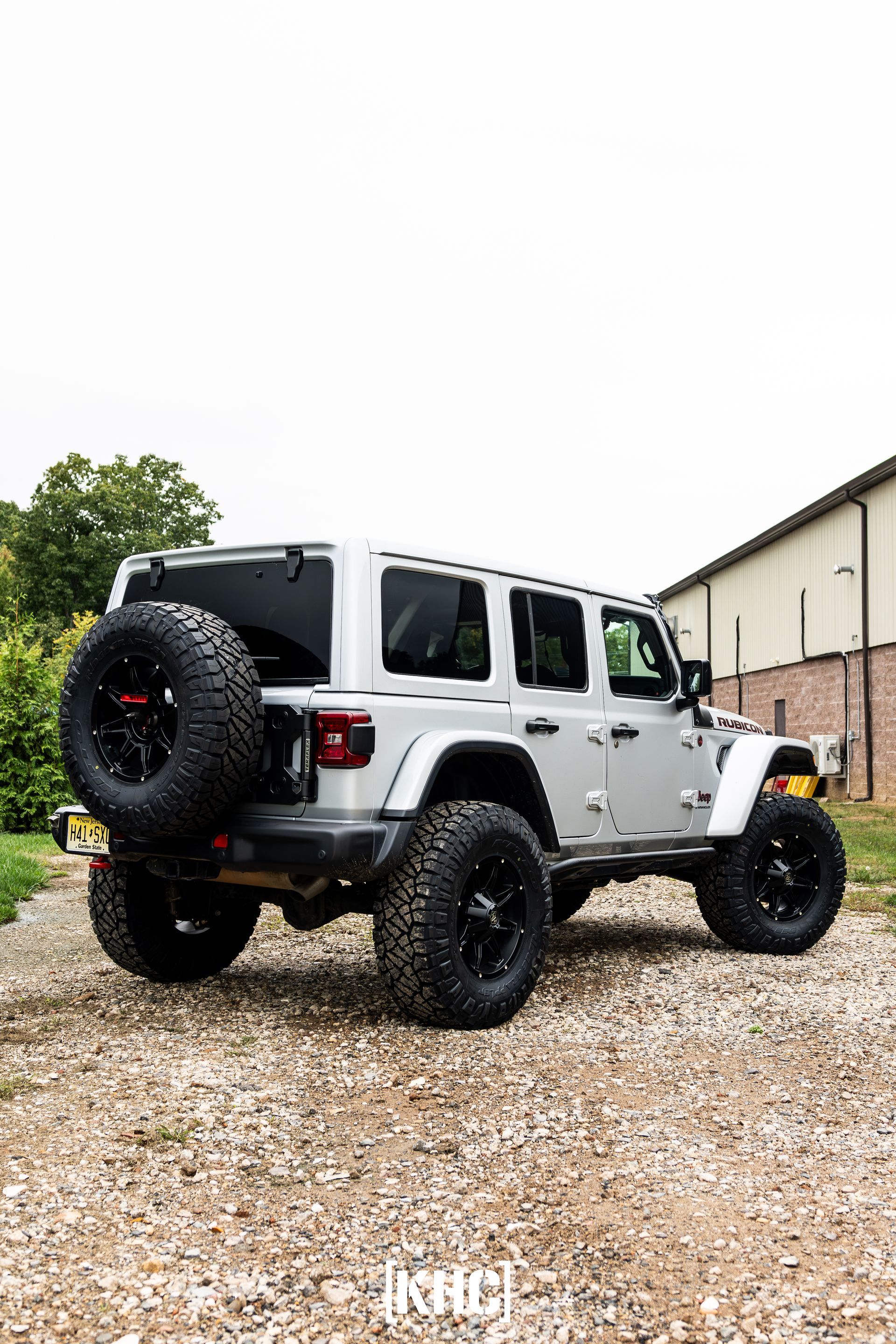 Silver Jeep Wrangler on gravel, large black tires, spare tire on the back, parked outdoors.