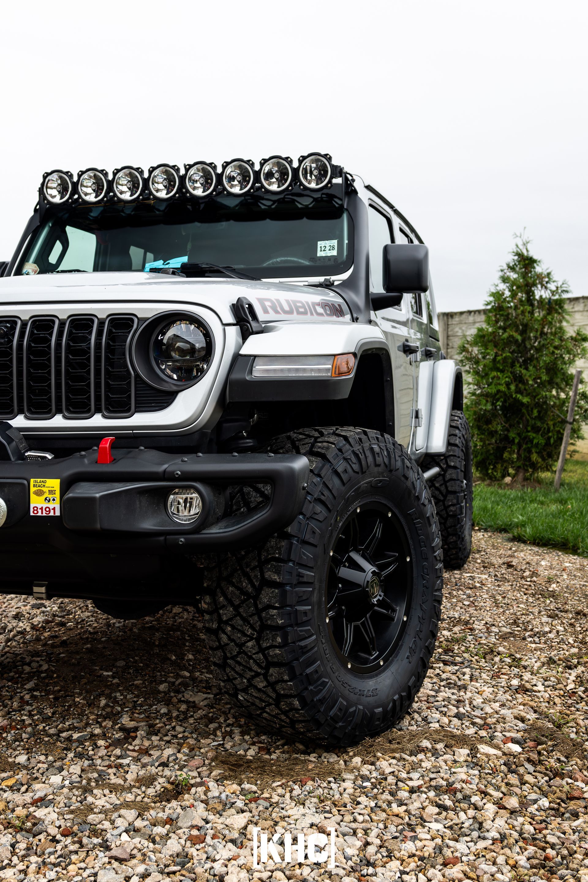 Silver Jeep with black accents, large tires, and roof lights parked on gravel.