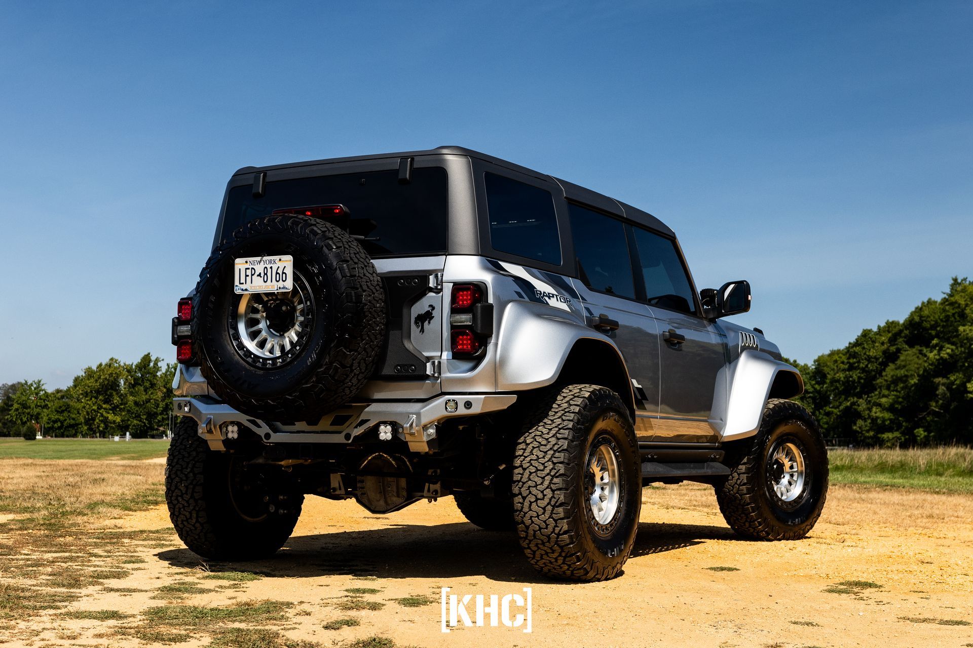 Silver Ford Bronco with black tires parked on dirt.