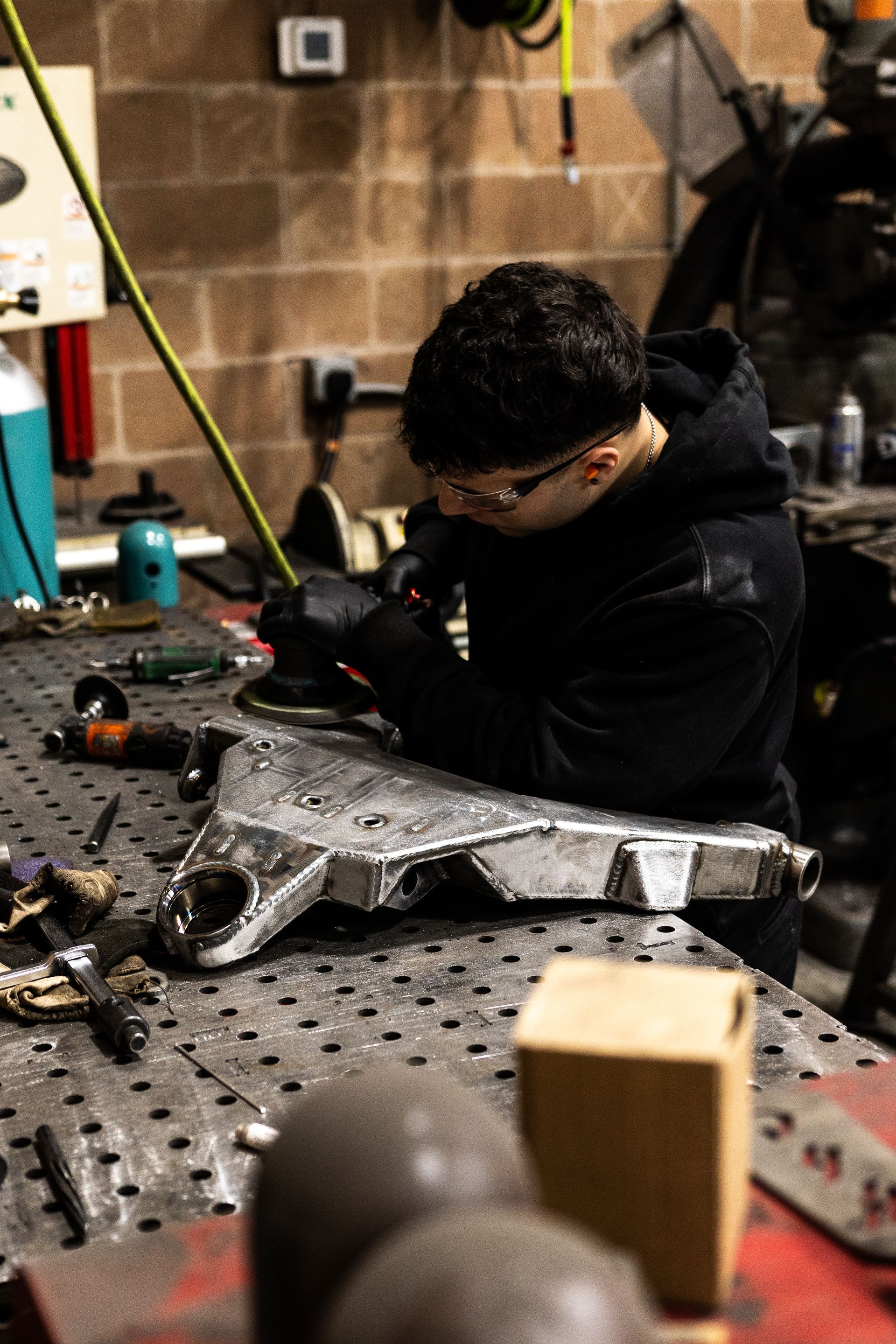Person grinds metal part at a workbench in a workshop. Wearing safety glasses and a black hoodie.