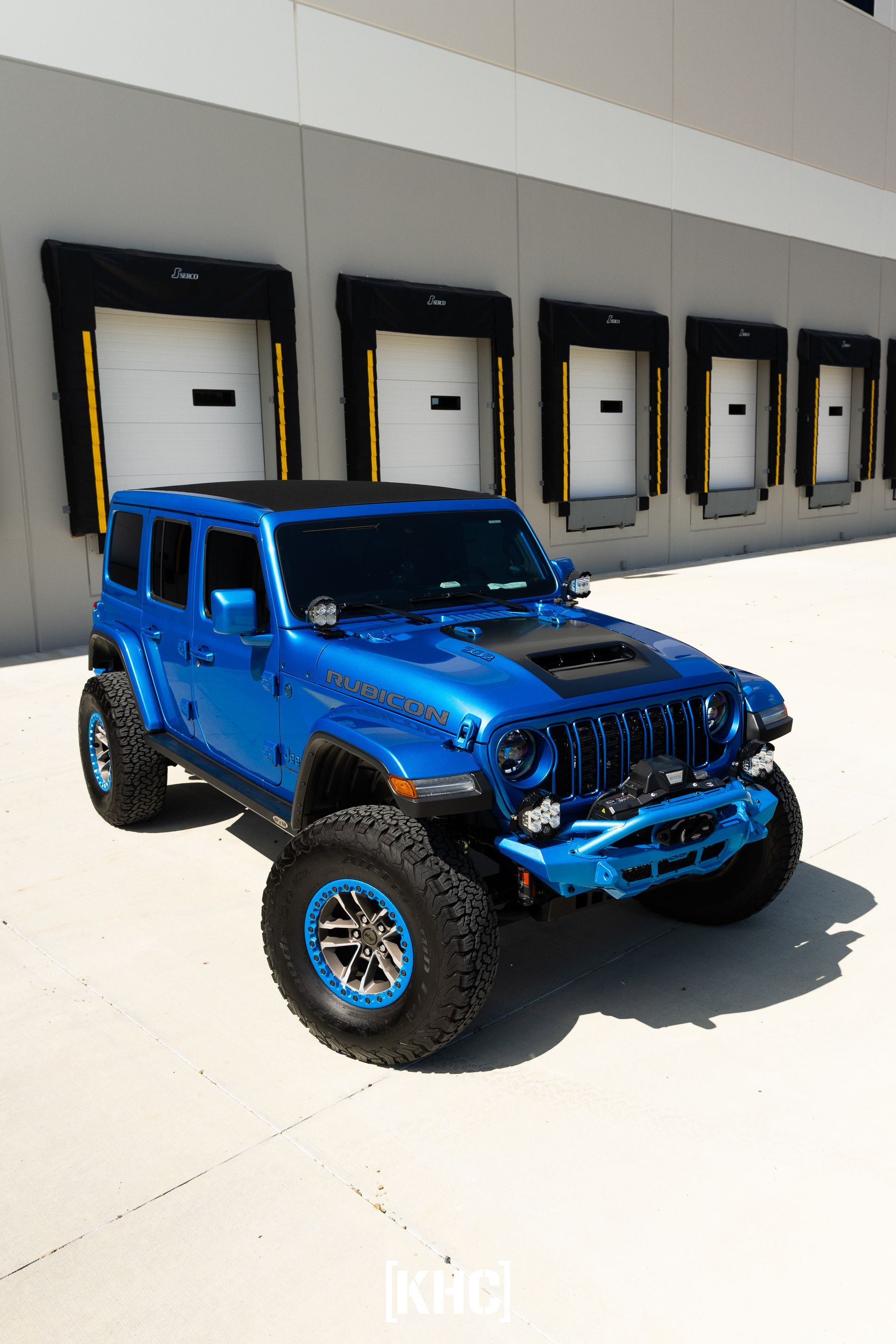 Blue Jeep Wrangler with off-road modifications, parked in front of loading docks.