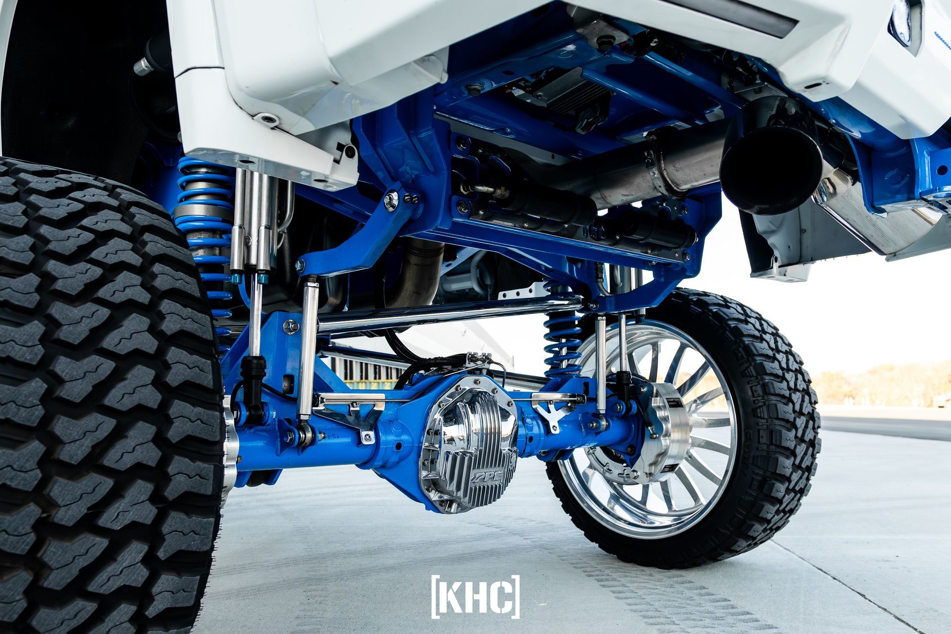 Blue lifted truck's undercarriage with chrome and oversized wheels on a paved surface.