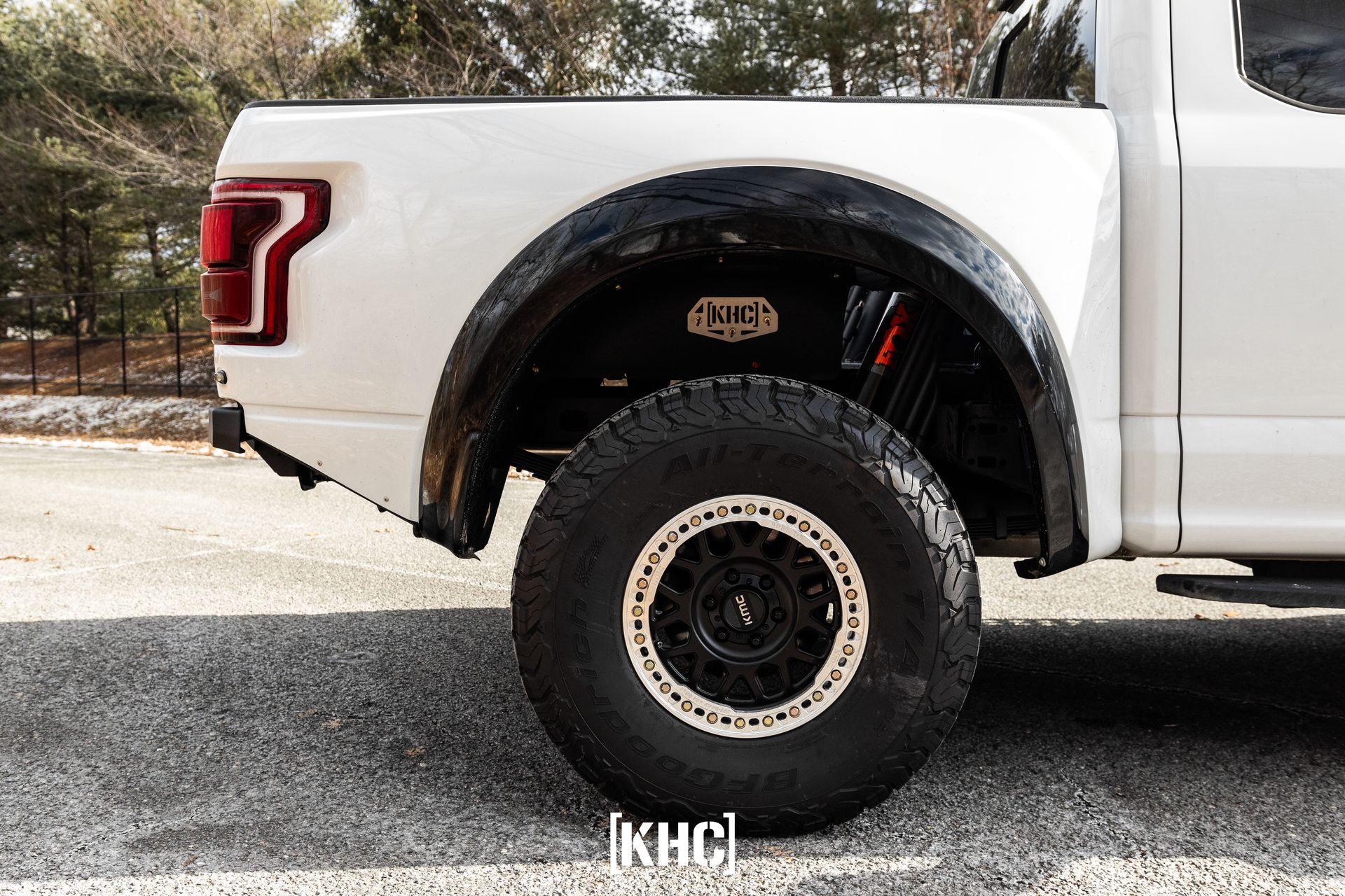 White truck with large off-road tire, black fender flare, and a red shock absorber on a paved road.