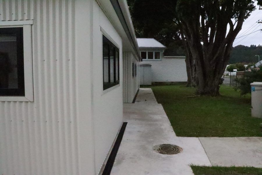 White house side walkway beside a grassy yard and large tree, with a concrete path and windows along the wall