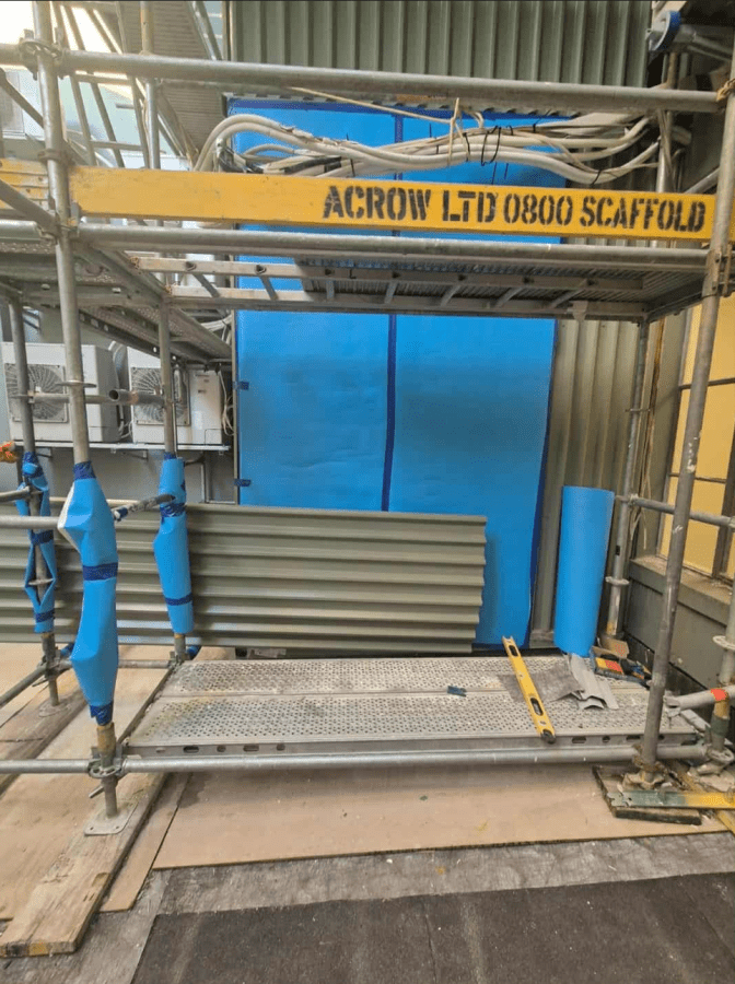 Scaffolded work area with blue tarp and stacked metal panels under an Acrow scaffold frame
