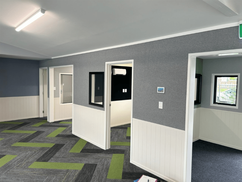 Empty office room with gray walls, patterned carpet, and white wainscoting, seen through an interior doorway