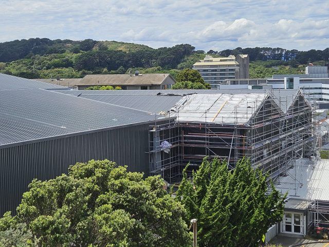 Greenhouse roof under construction beside a large industrial building, with trees and hills in the background