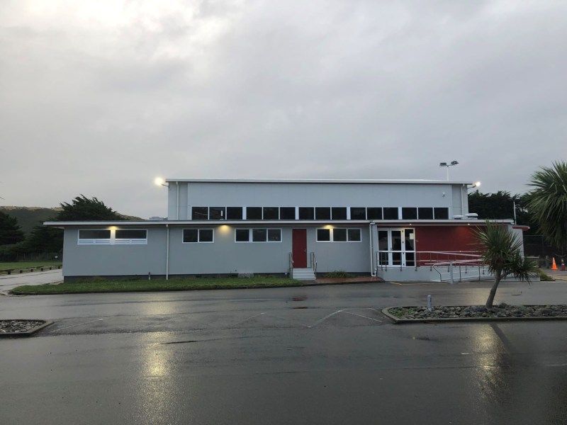 Low, modern white building with red doors at dusk, reflected on a wet road under a cloudy sky